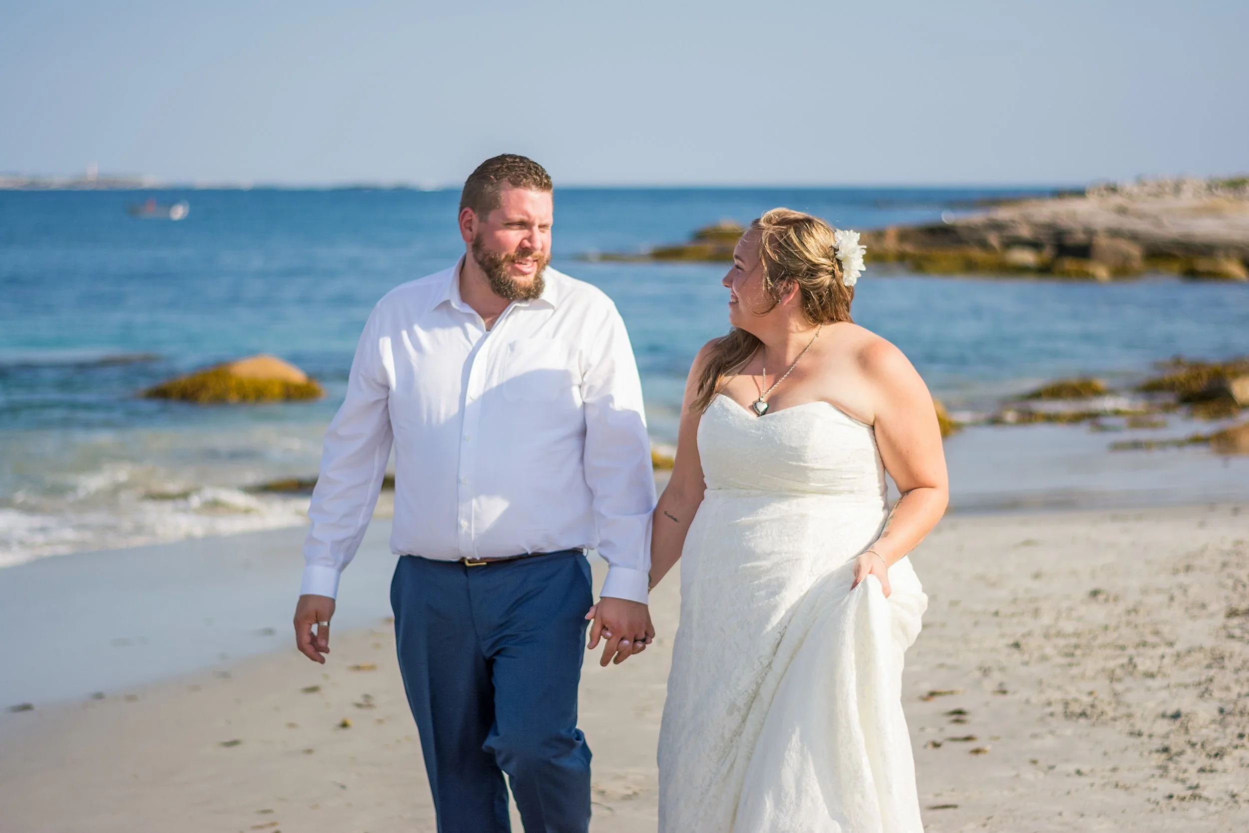 A couple in wedding attire holding hands and walking on a beach with rocks, ocean, and clear blue sky in the background.