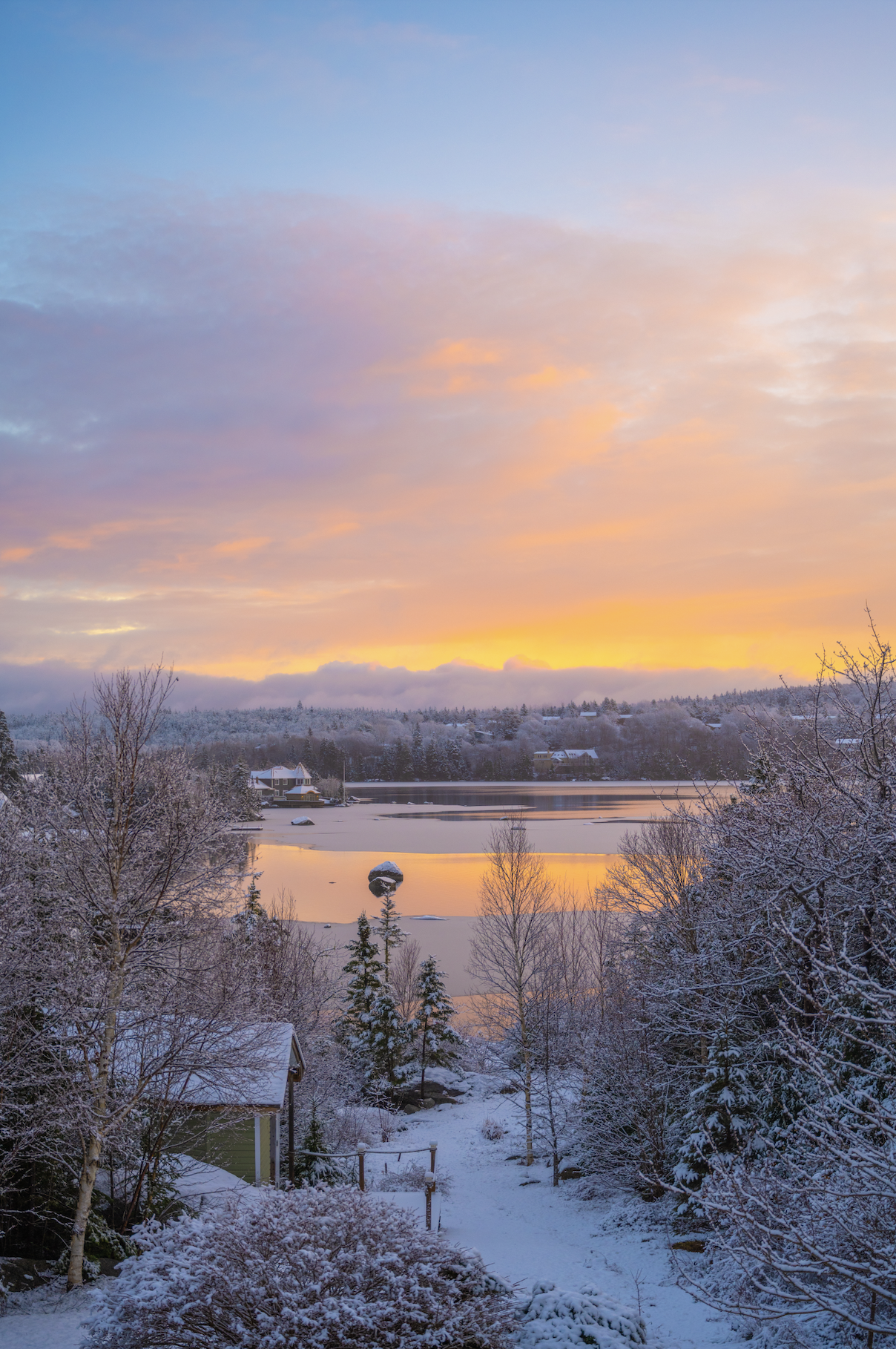 Snow-covered landscape with trees, houses, and a partially frozen lake reflecting colors of a sunset or sunrise, with a colorful sky overhead.