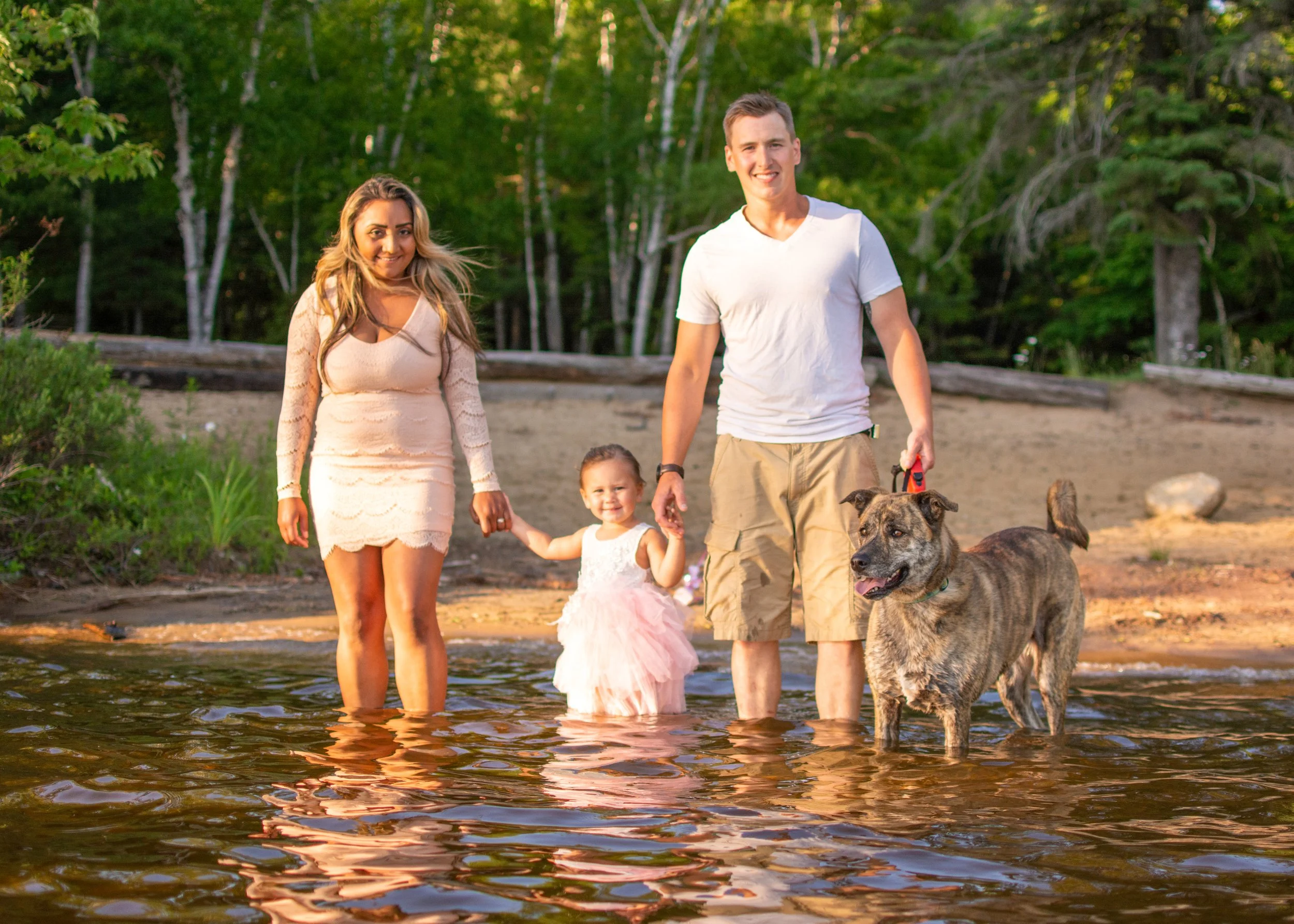A family of four, including a woman, a man, a young girl, and a dog, standing in shallow water at a lakeshore with trees in the background during sunset.