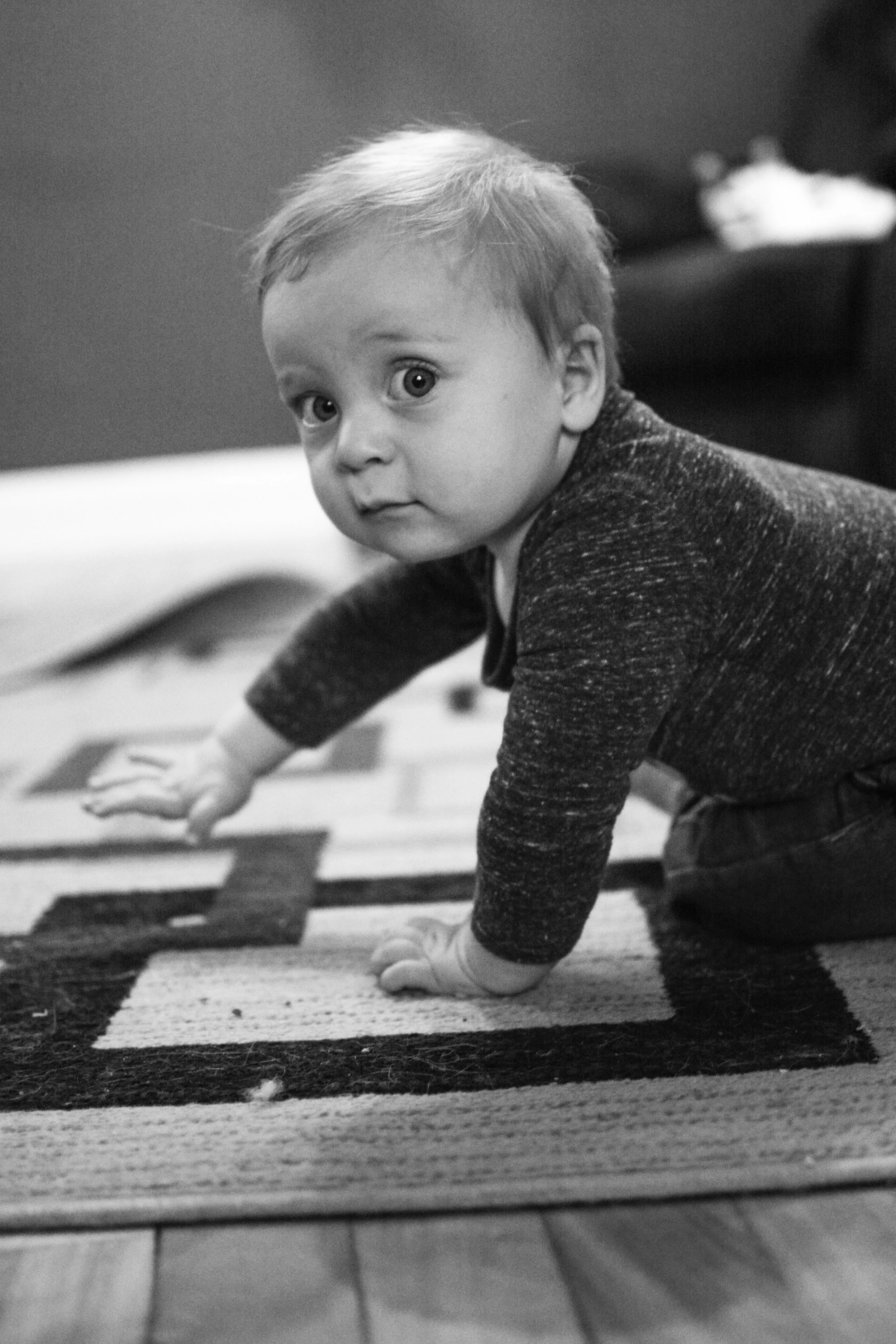 A young child is crawling on a patterned rug, looking into the camera with wide eyes.