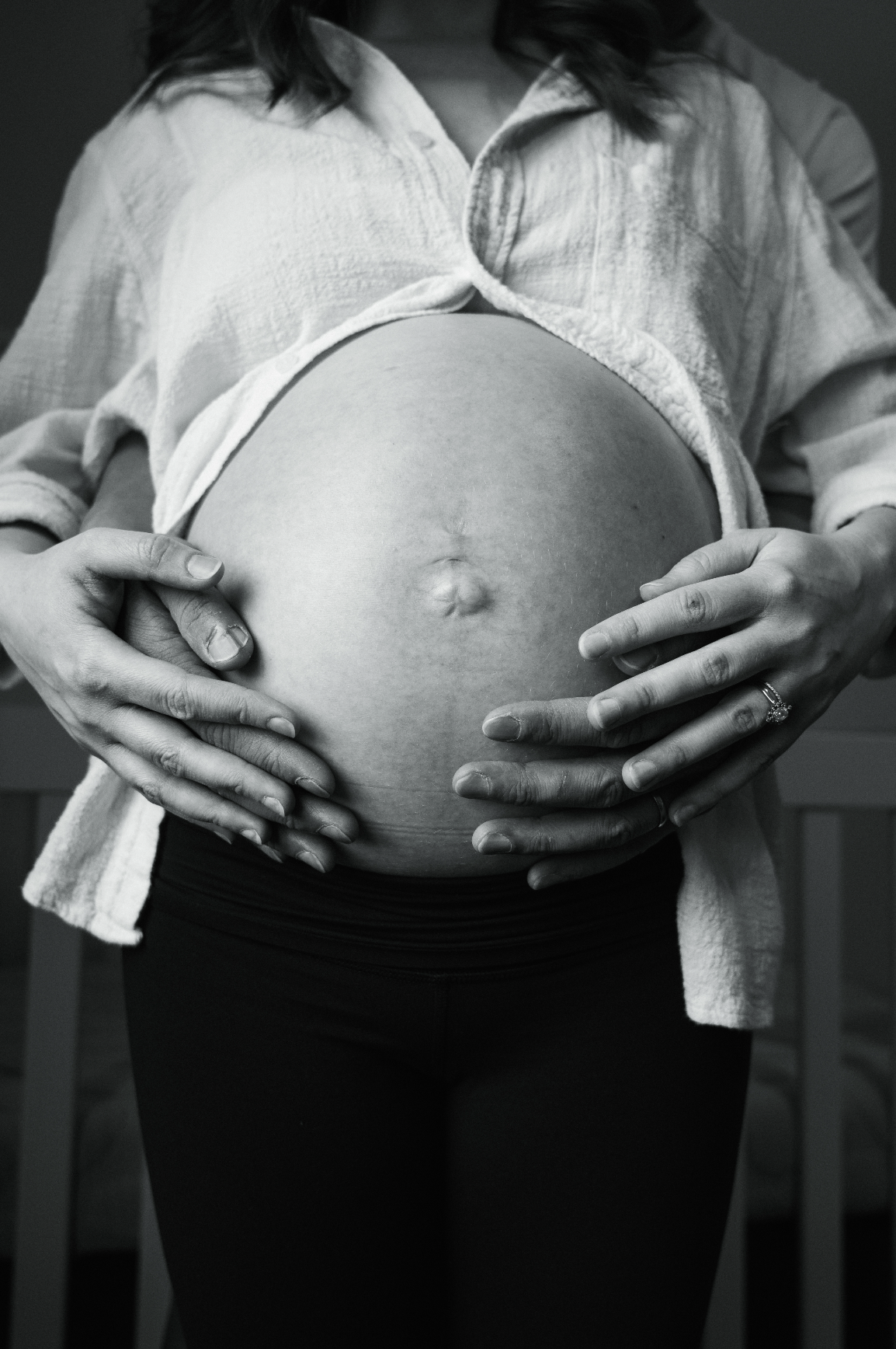 A pregnant woman holding her belly with both hands, wearing a partially unbuttoned shirt and black leggings. The image is in black and white.