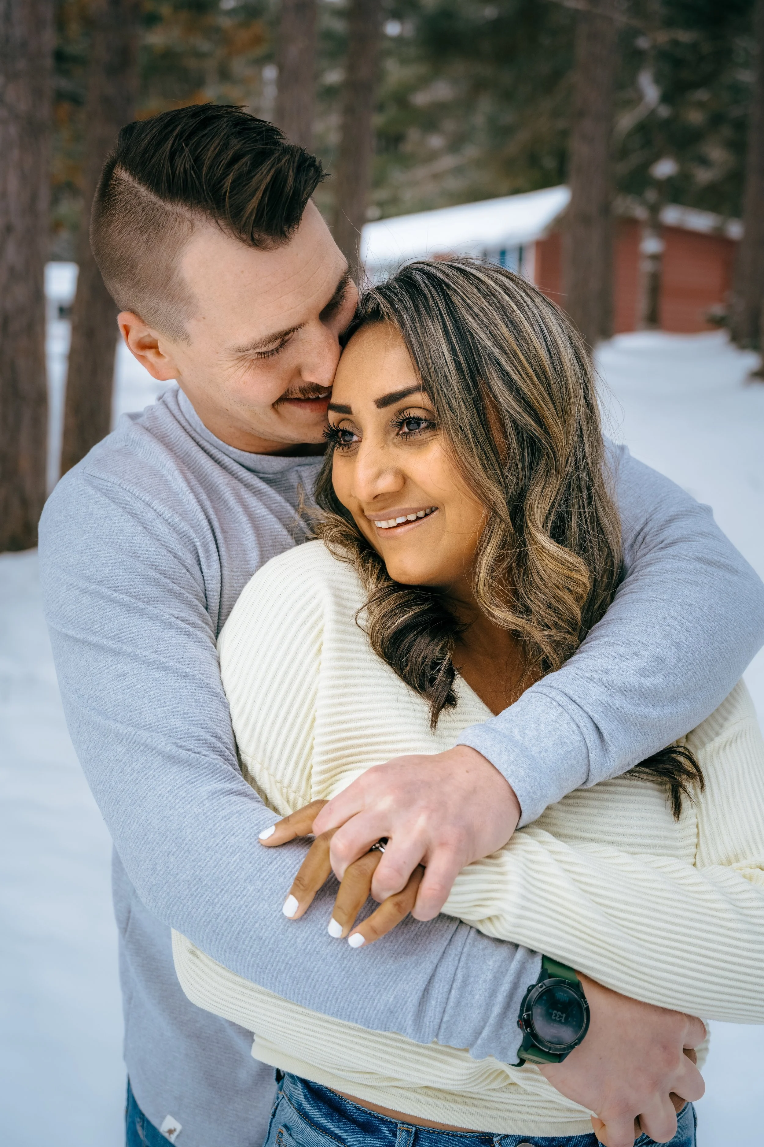 A young couple embraces outdoors in a snowy setting with trees in the background.