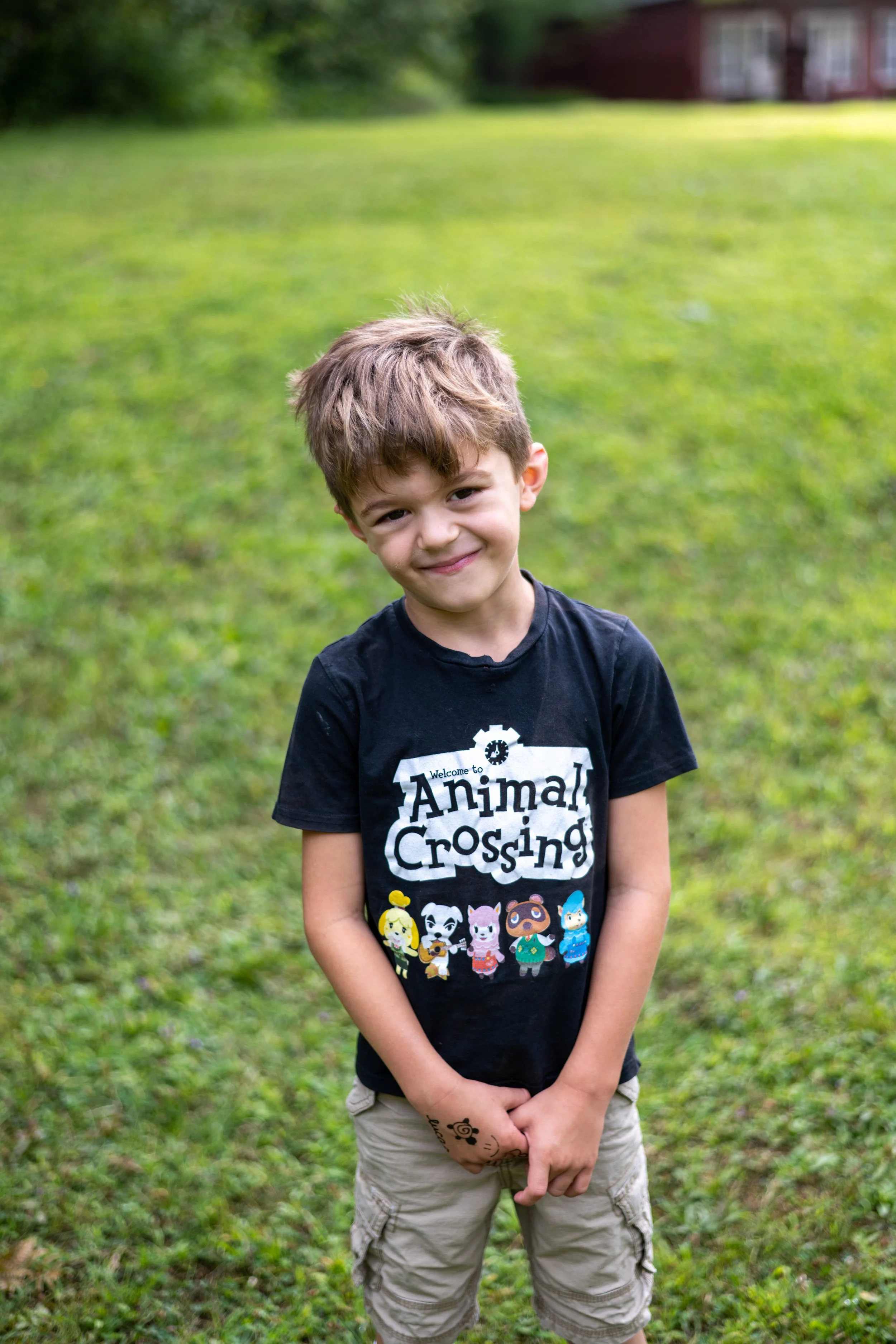 A young boy with brown hair smiling, wearing a black Animal Crossing t-shirt and beige cargo shorts, standing on a grassy field outdoors.