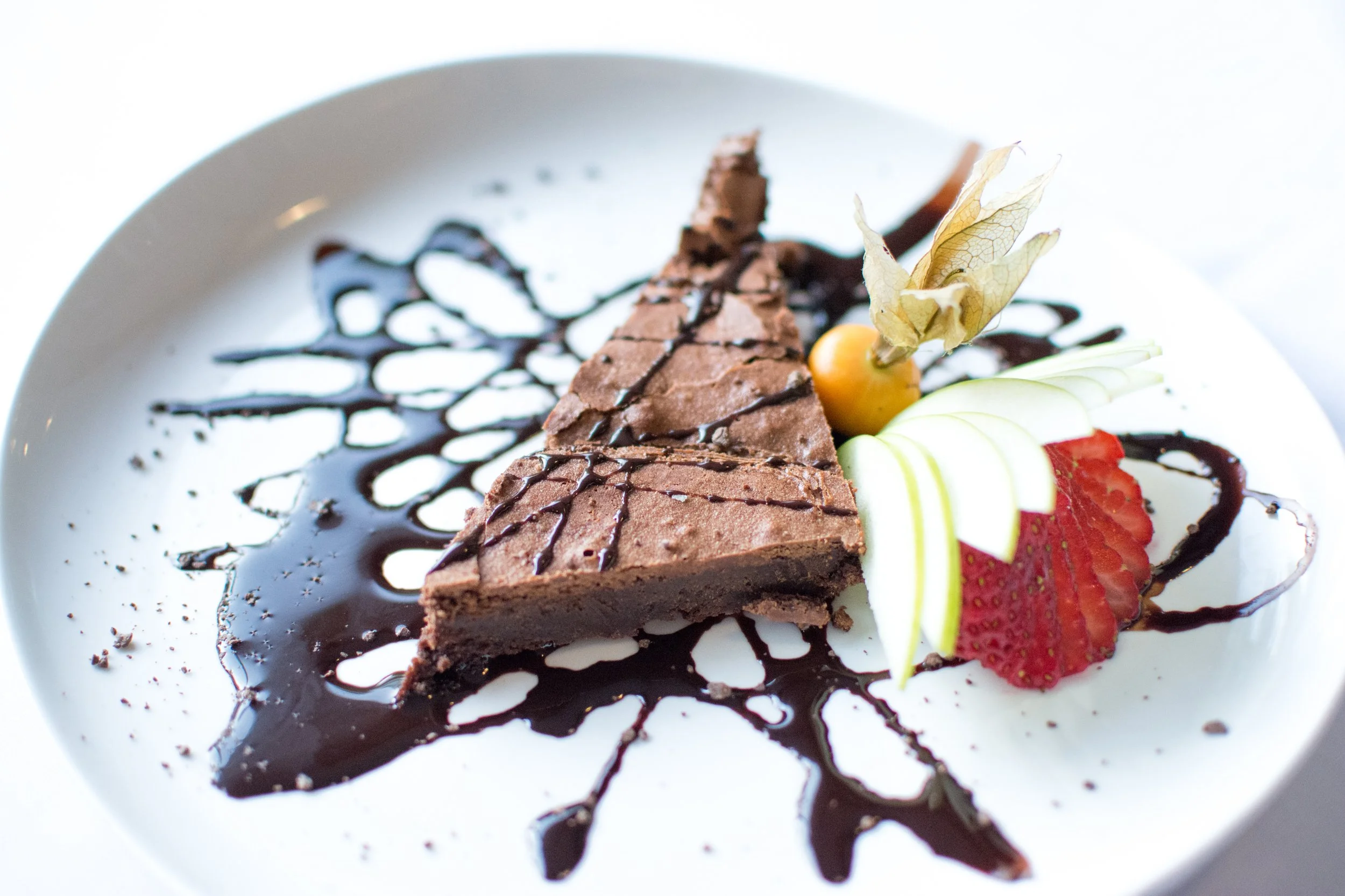 Chocolate tart with chocolate drizzle, sliced strawberries, apple slices, a cape gooseberry, and a physalis, garnished on a white plate with chocolate sauce design.