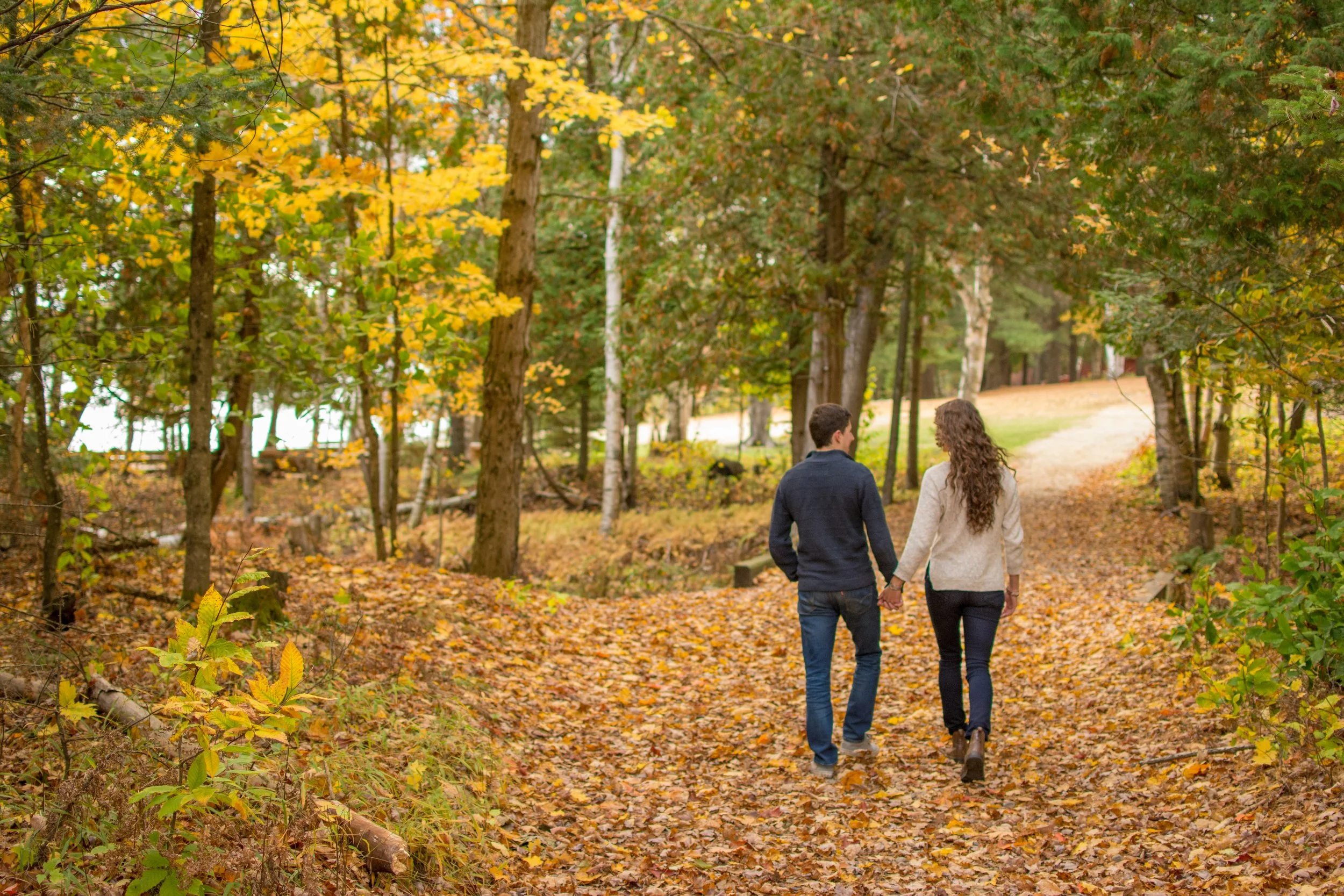 A young couple walking hand in hand through a wooded park during autumn with fallen leaves on the ground.