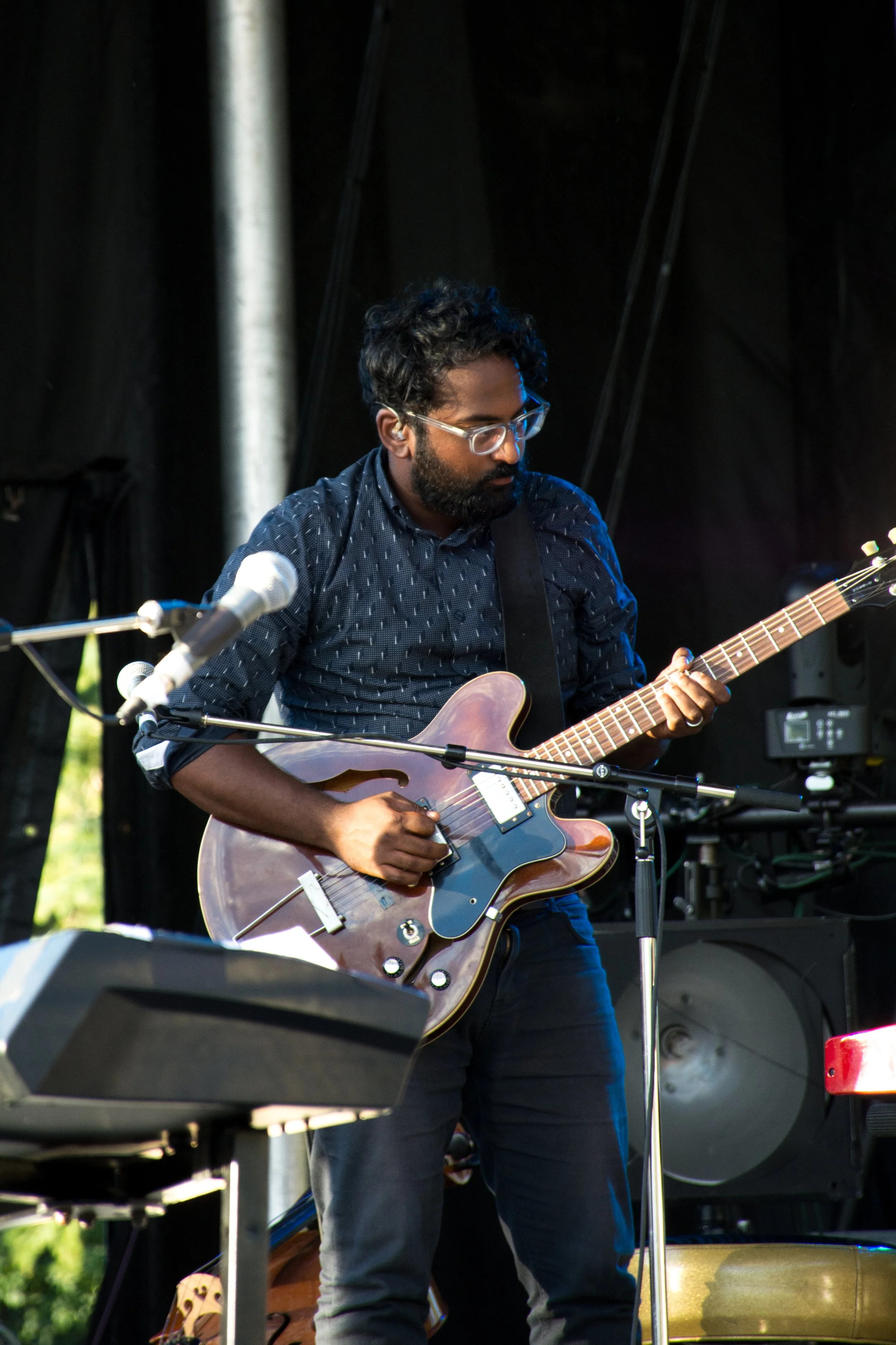 A man with glasses and a beard playing an electric guitar on stage at an outdoor concert.