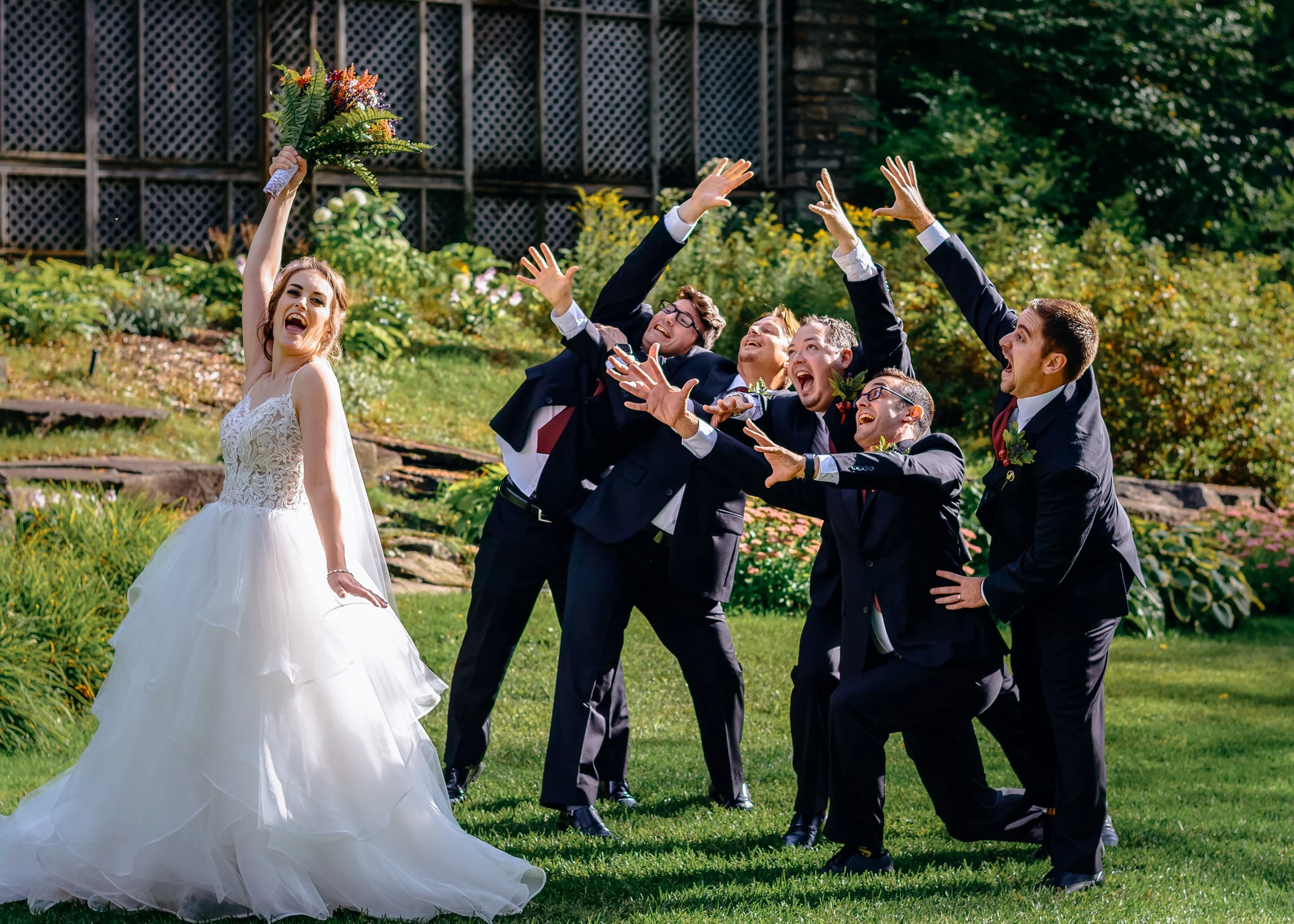A bride in a white wedding gown holding a bouquet of flowers and smiling, surrounded by five groomsmen in dark suits, as they pose playfully outdoors on a sunny day with greenery and flowers in the background.