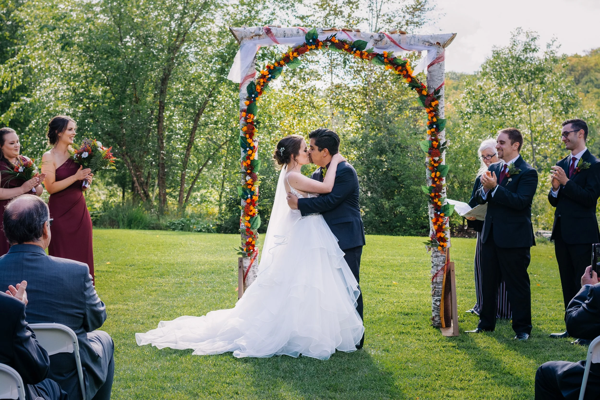 A wedding ceremony outdoors with a bride and groom kissing under a floral arch, surrounded by bridesmaids and groomsmen clapping and holding bouquets on a grassy lawn with trees in the background.