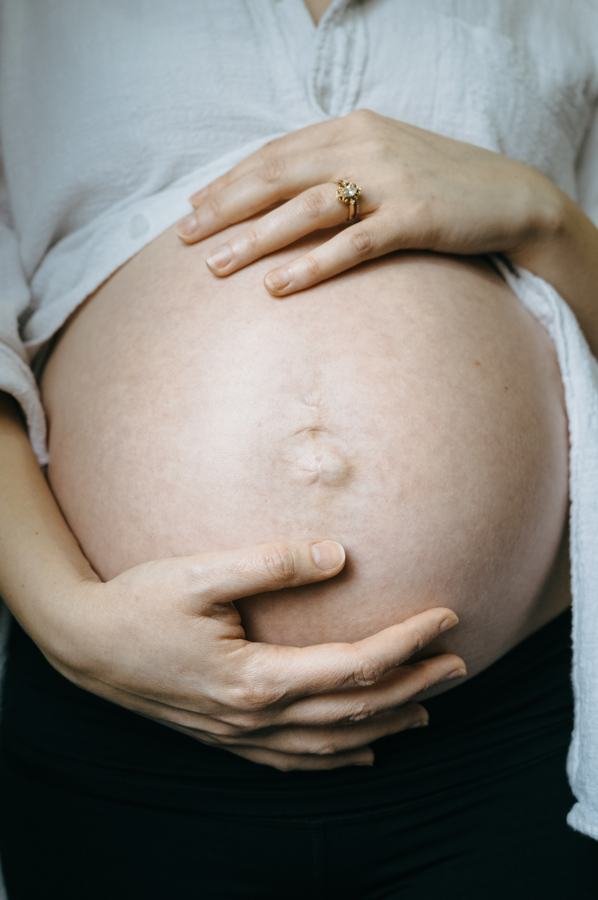 Close-up of a pregnant woman’s belly, with hands gently cradling it. The woman is wearing a white shirt and a ring on her finger.