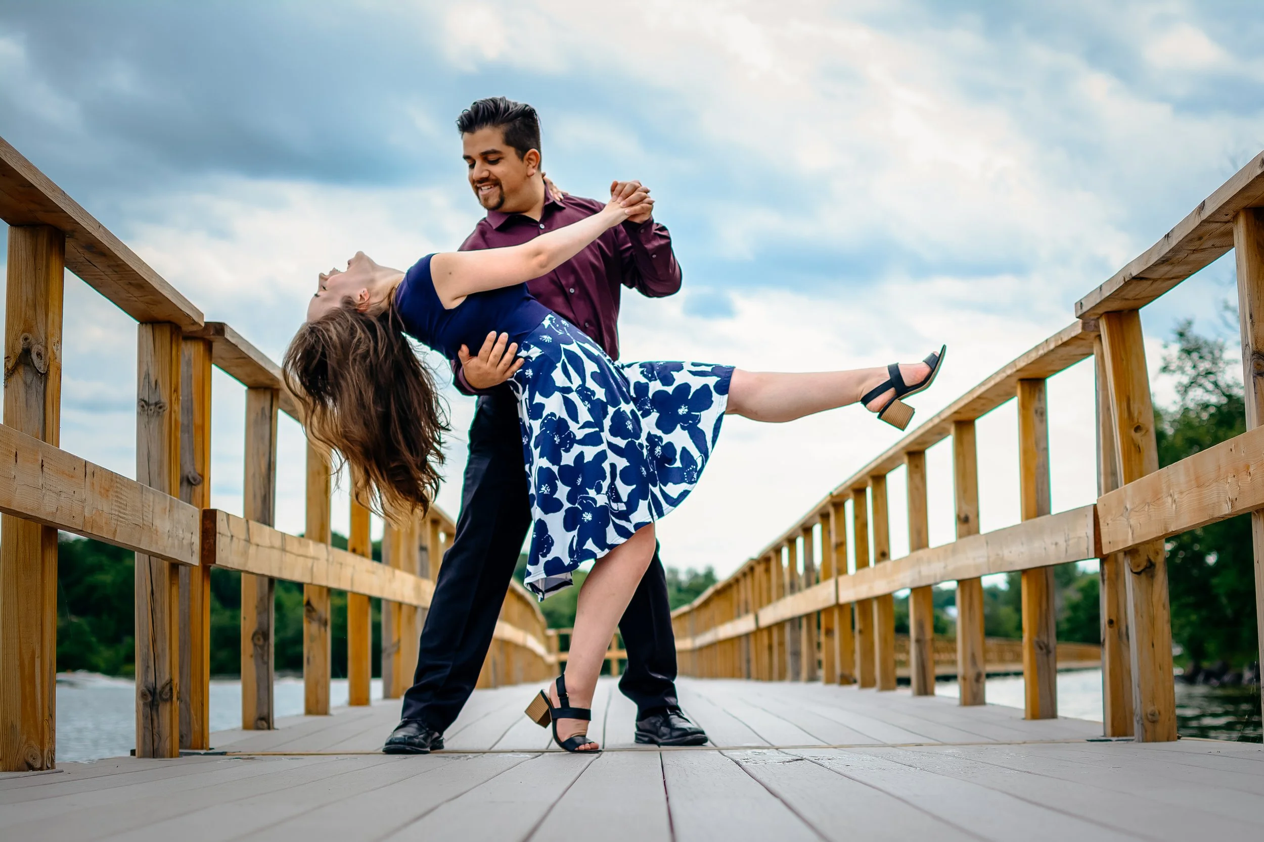 A man and a woman dancing on a wooden bridge over water, with the man holding the woman as she leans back and extends one leg.