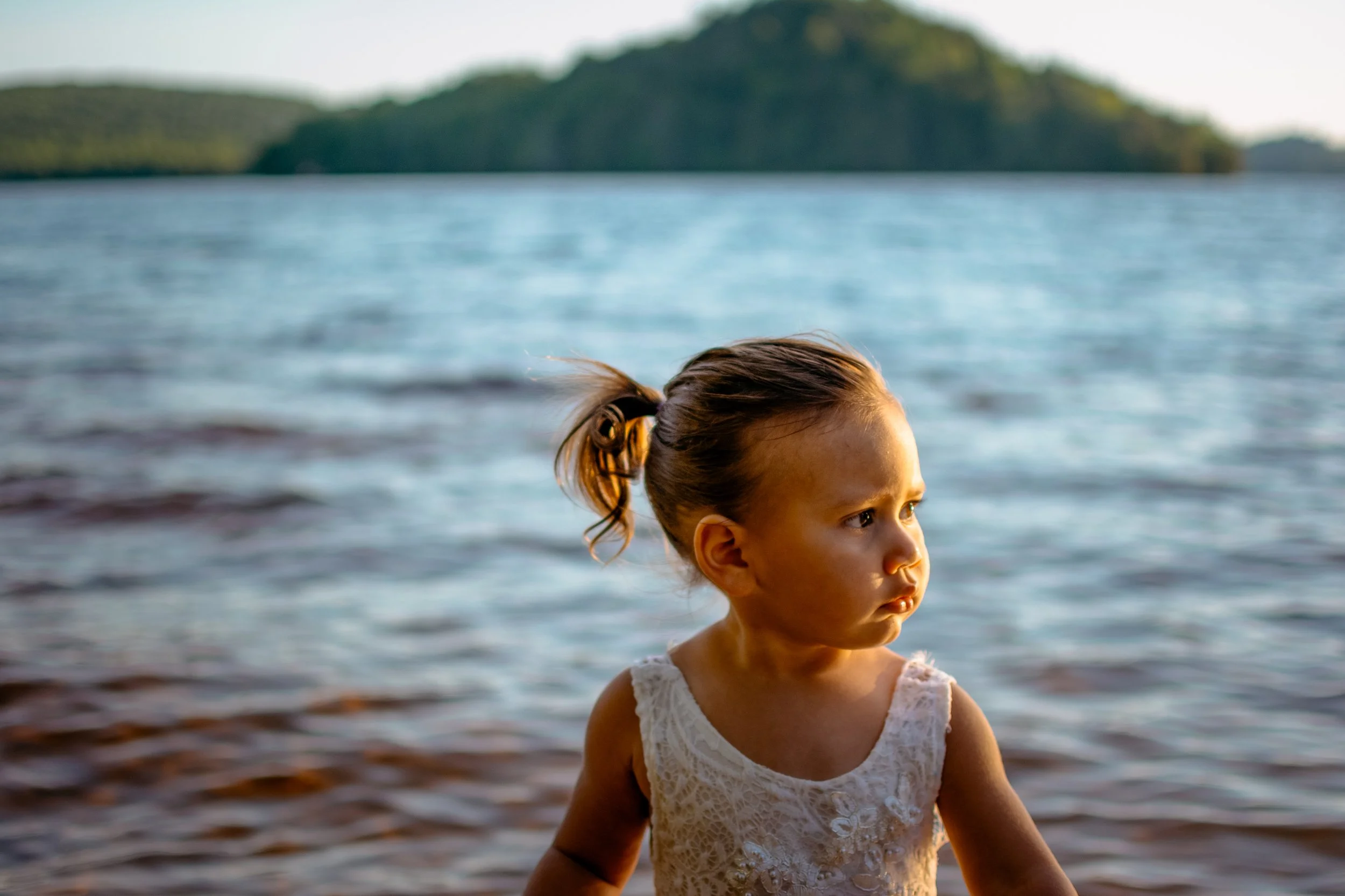 A young girl with a ponytail wearing a white lace dress standing near a body of water with trees in the background.