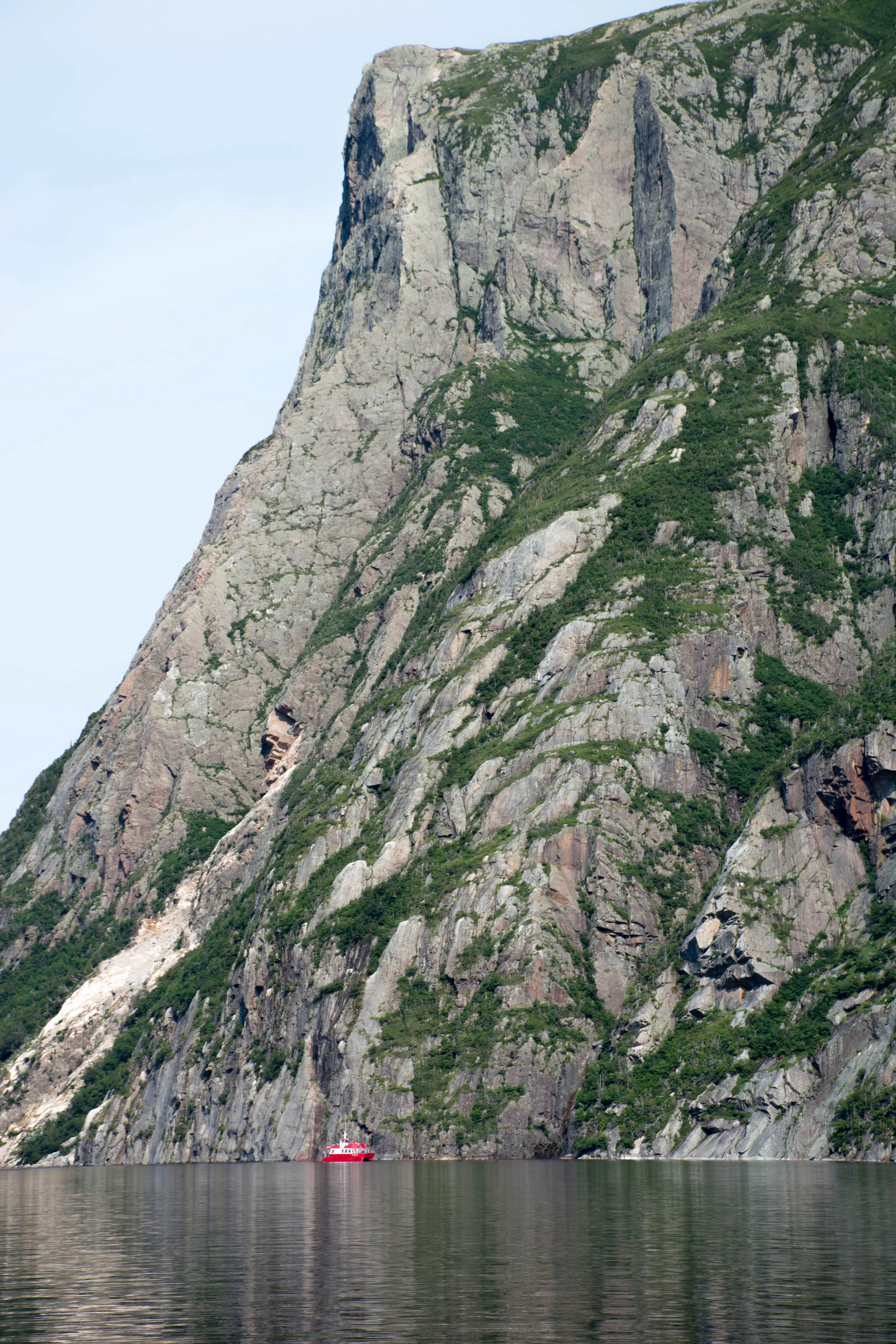 A large steep rock cliff beside calm water with a small red boat near the base.