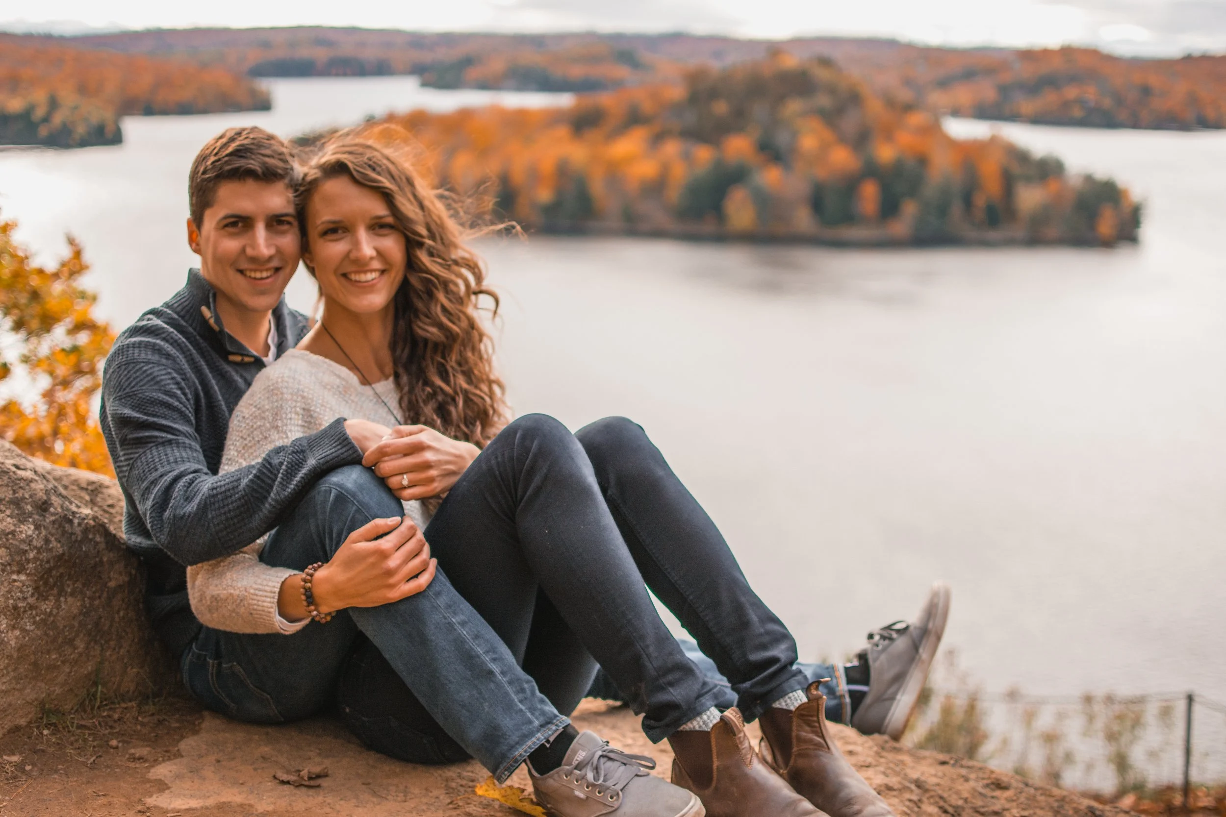 A smiling couple sitting on a rock with a lake and autumn-colored trees in the background.