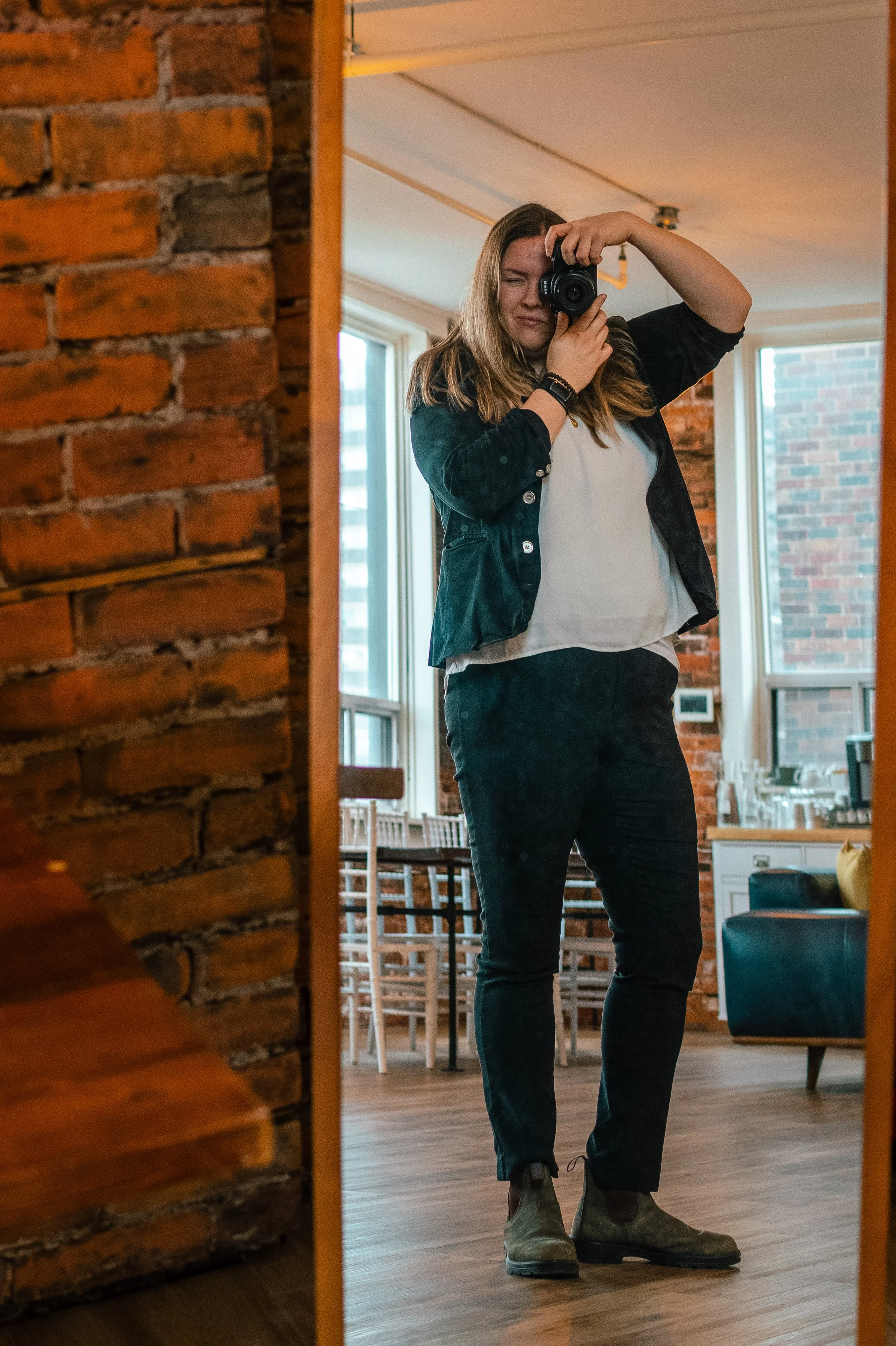 A woman taking a selfie in a mirror in a room with exposed brick walls, large windows, and wooden flooring.