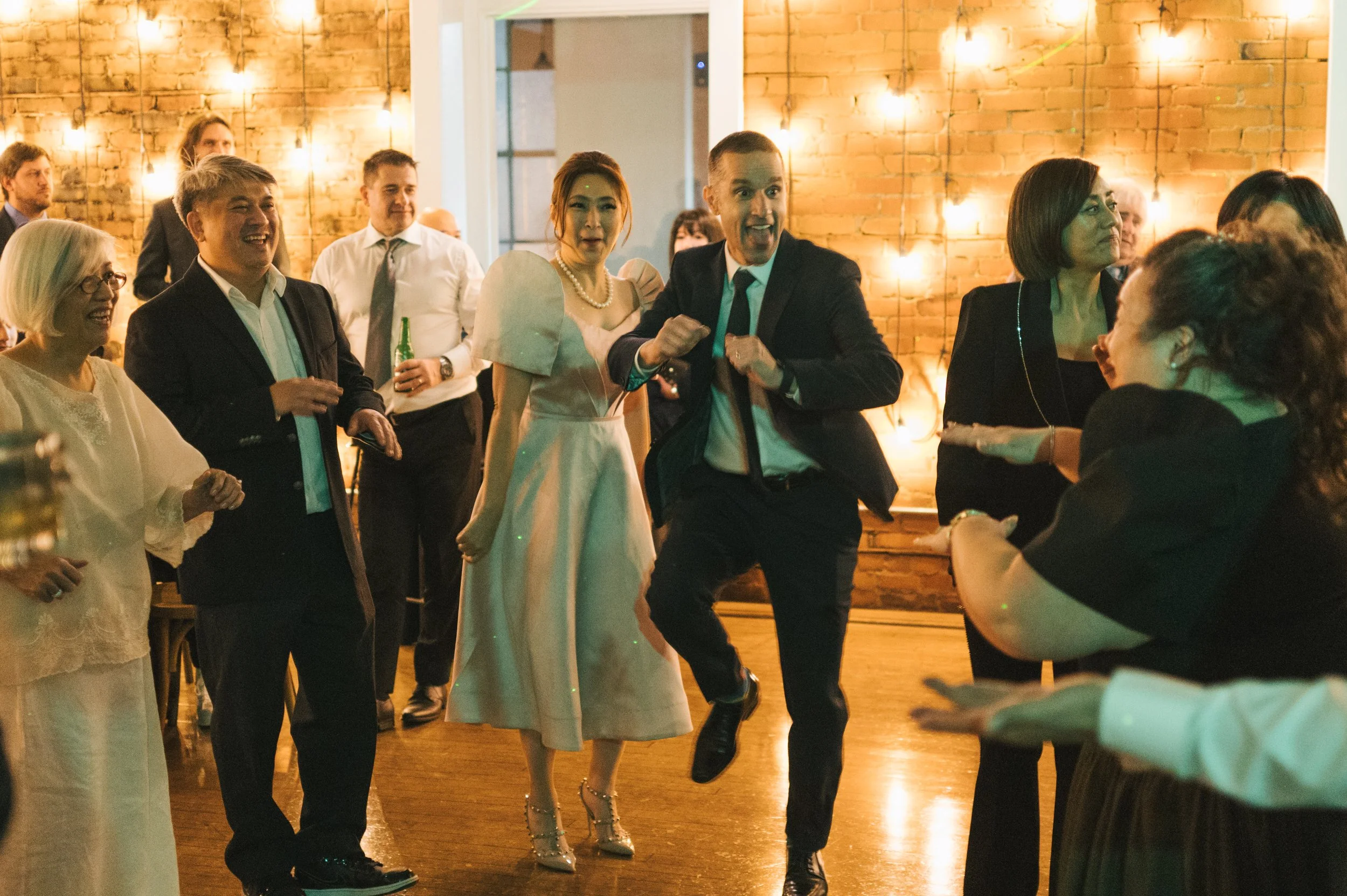 People dancing and socializing at a party in a warmly lit room with brick walls and string lights.
