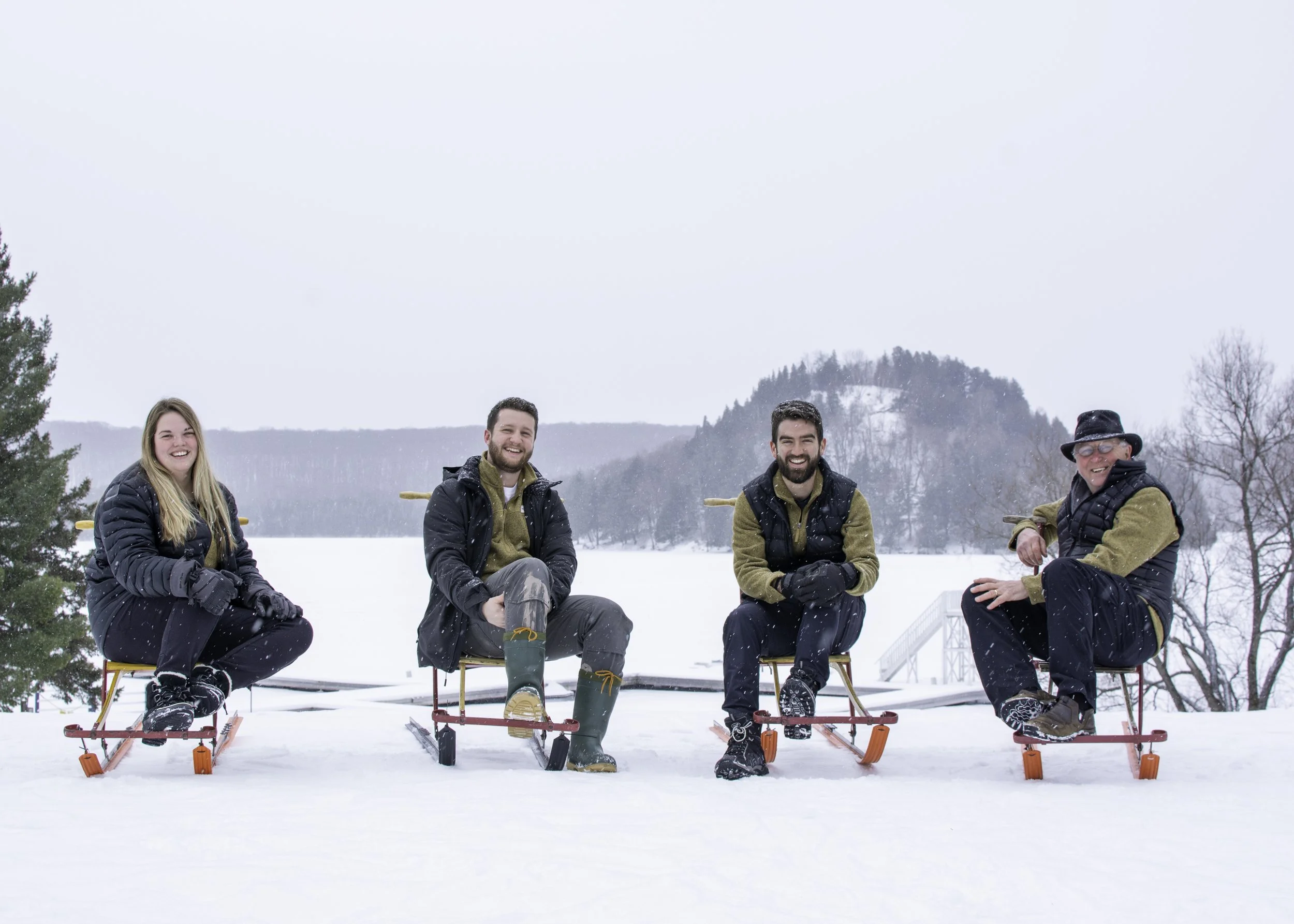 Four people sitting on sleds in a snowy landscape with a wooded hill in the background, smiling and enjoying winter.