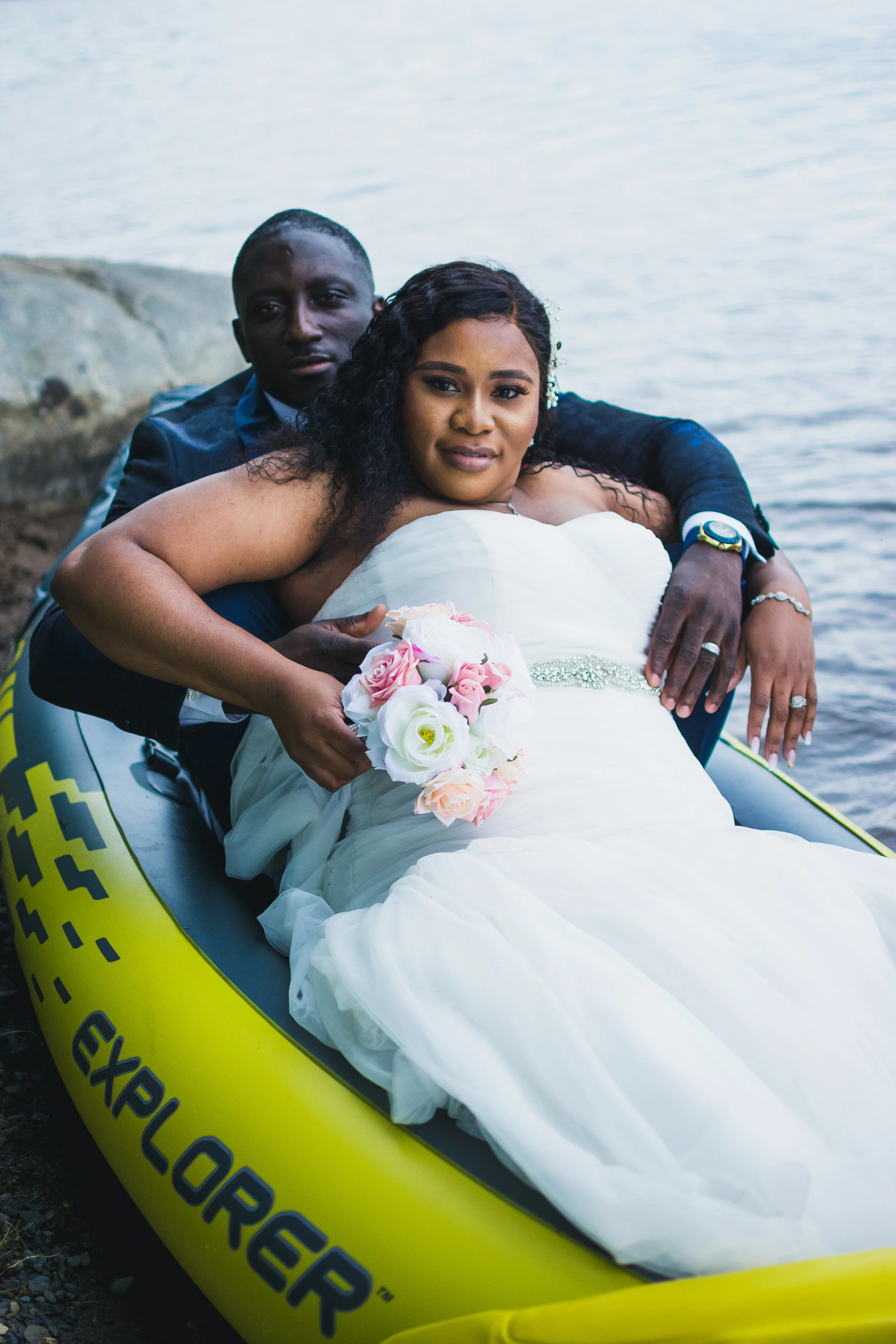 Couple in wedding attire sitting in an inflatable boat by water, with the bride holding a bouquet of pink and white roses.