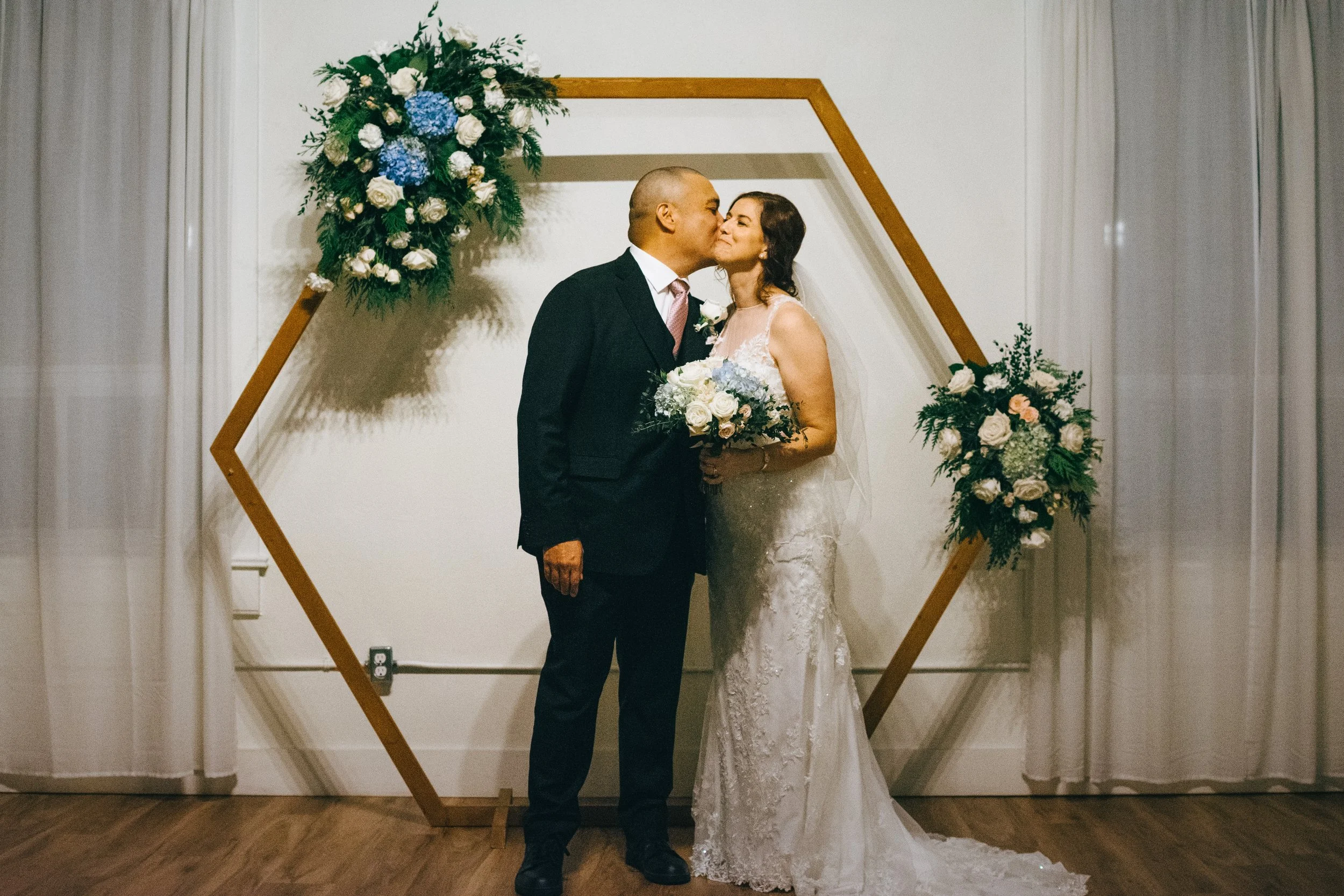 A newlywed couple sharing a kiss during their wedding ceremony, standing in front of a geometric floral arch decorated with white and blue flowers and green foliage.