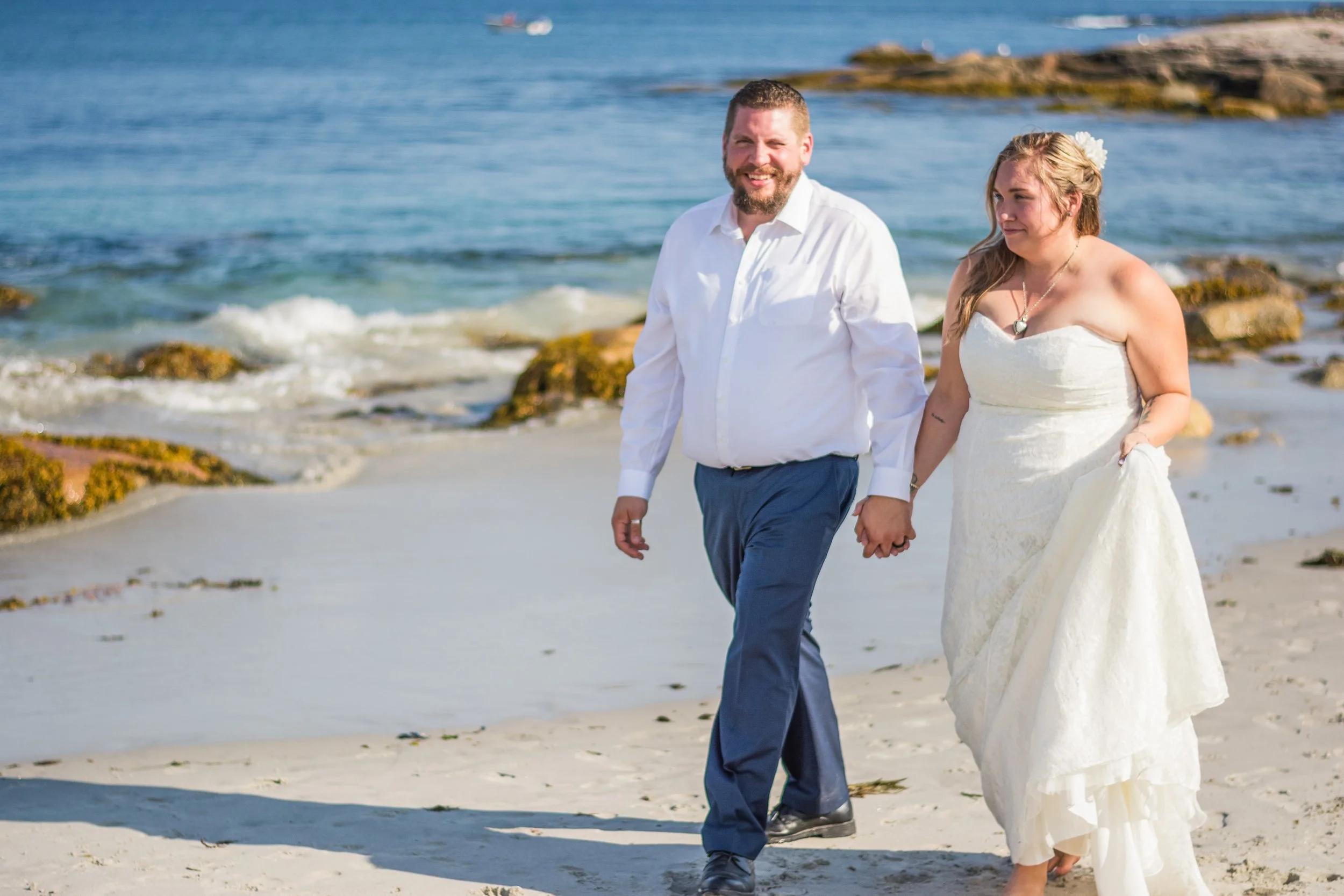 A couple in wedding attire walking hand-in-hand on a beach with rocks and ocean in the background.