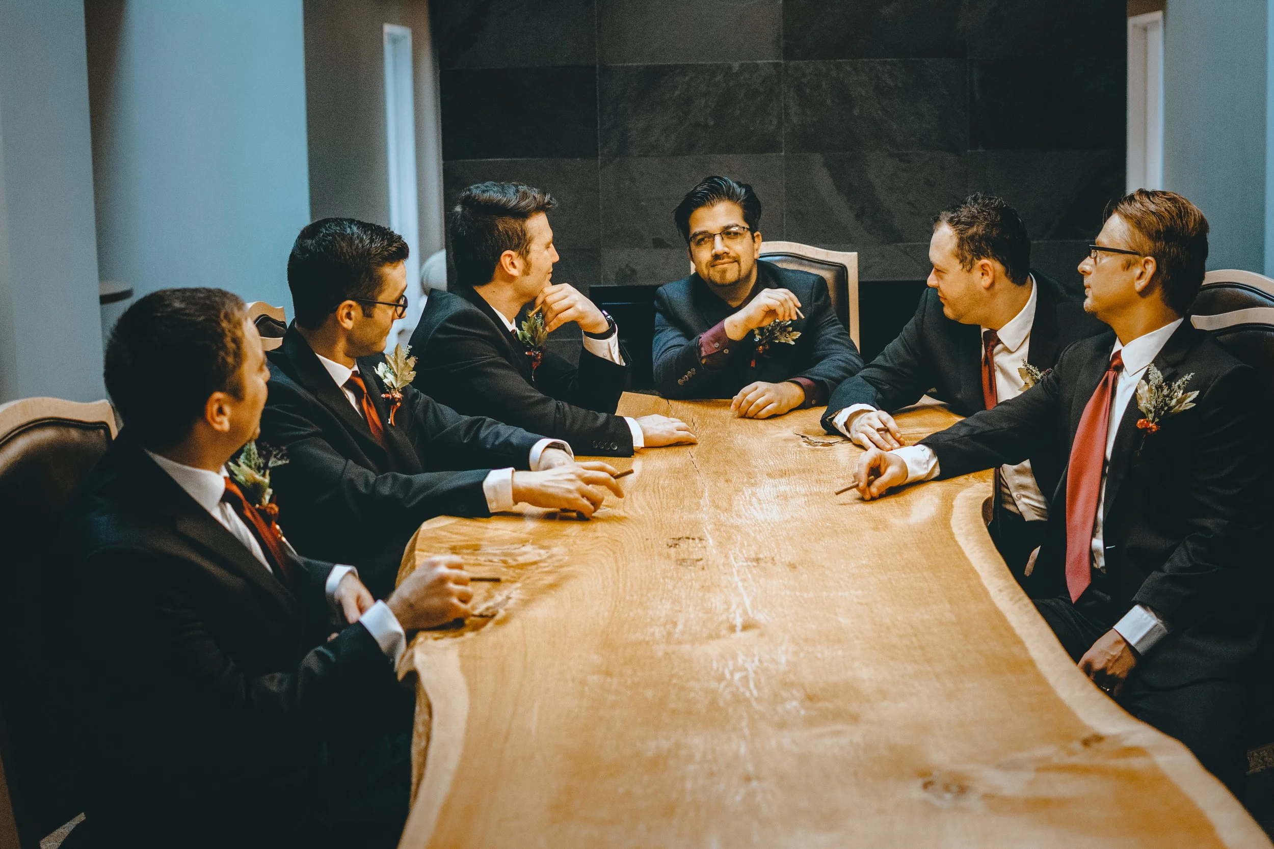A group of seven men in suits and ties sitting around a wooden conference table in a formal meeting room, engaged in conversation.