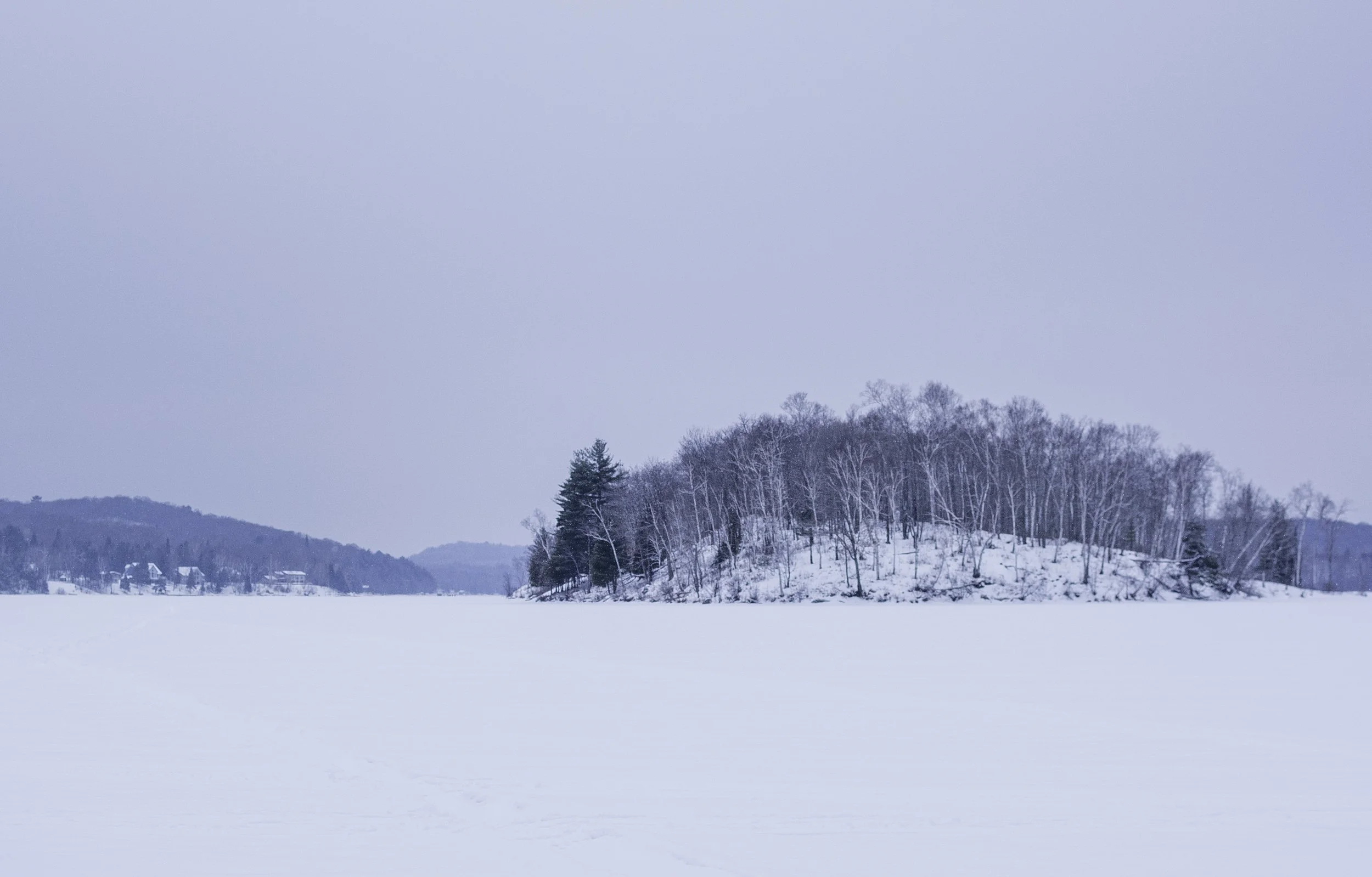 Frozen lake with snow-covered trees and hills in the background under a cloudy sky.