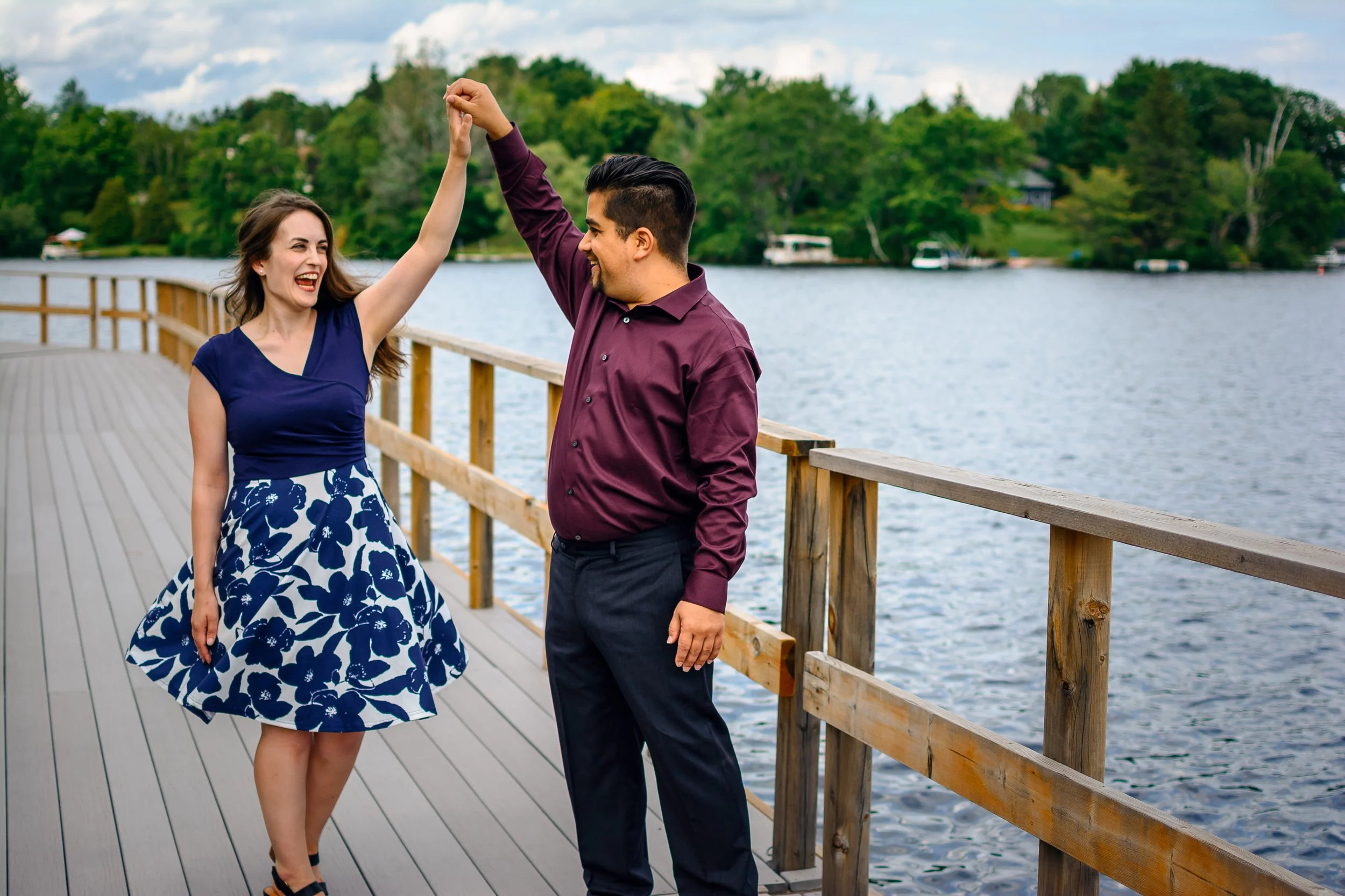 A man and woman dancing on a wooden pier by a lake with trees in the background, woman wearing a blue floral dress and man wearing a maroon shirt.