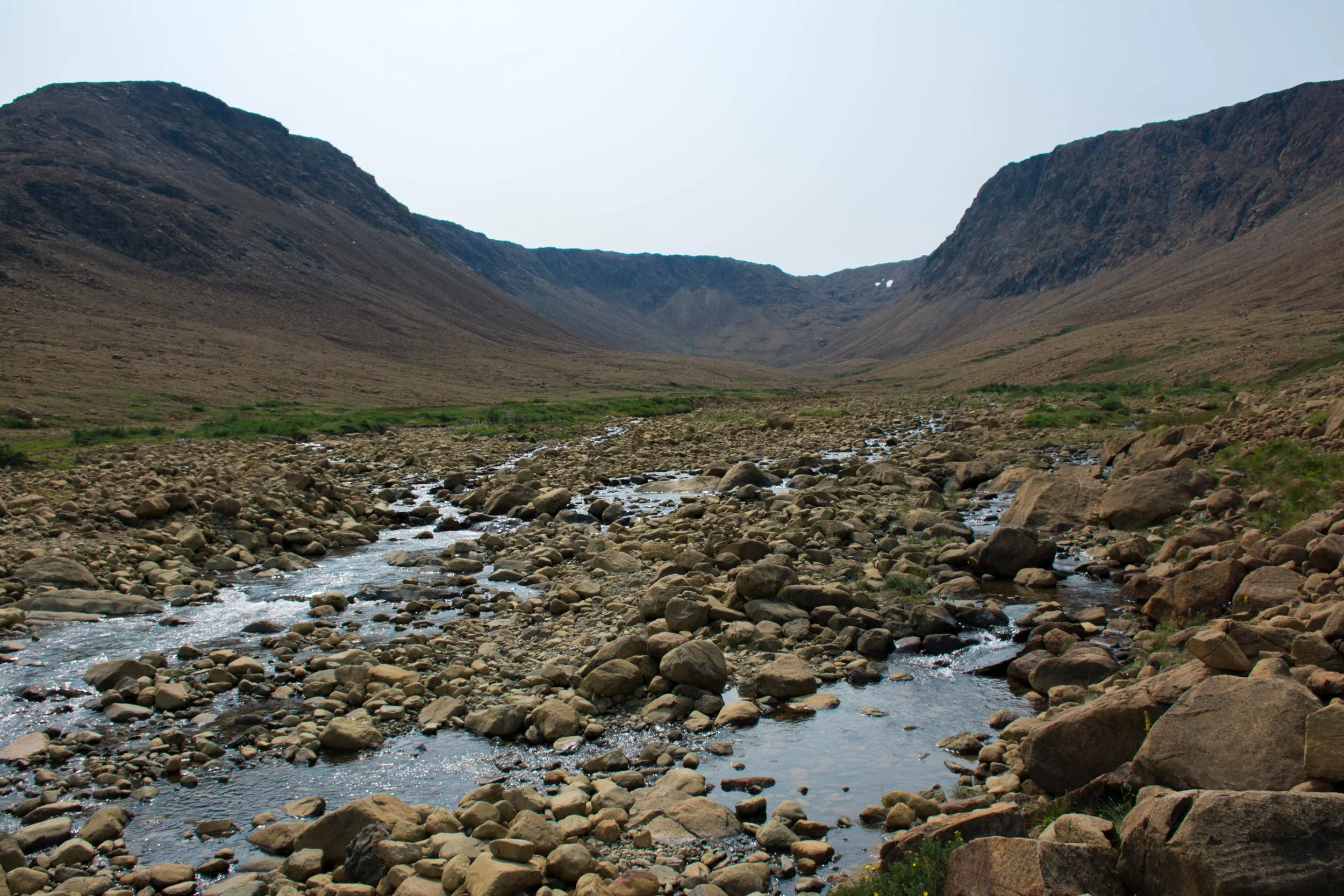 A mountain valley with a rocky stream running through it, surrounded by bare hills under a cloudy sky.