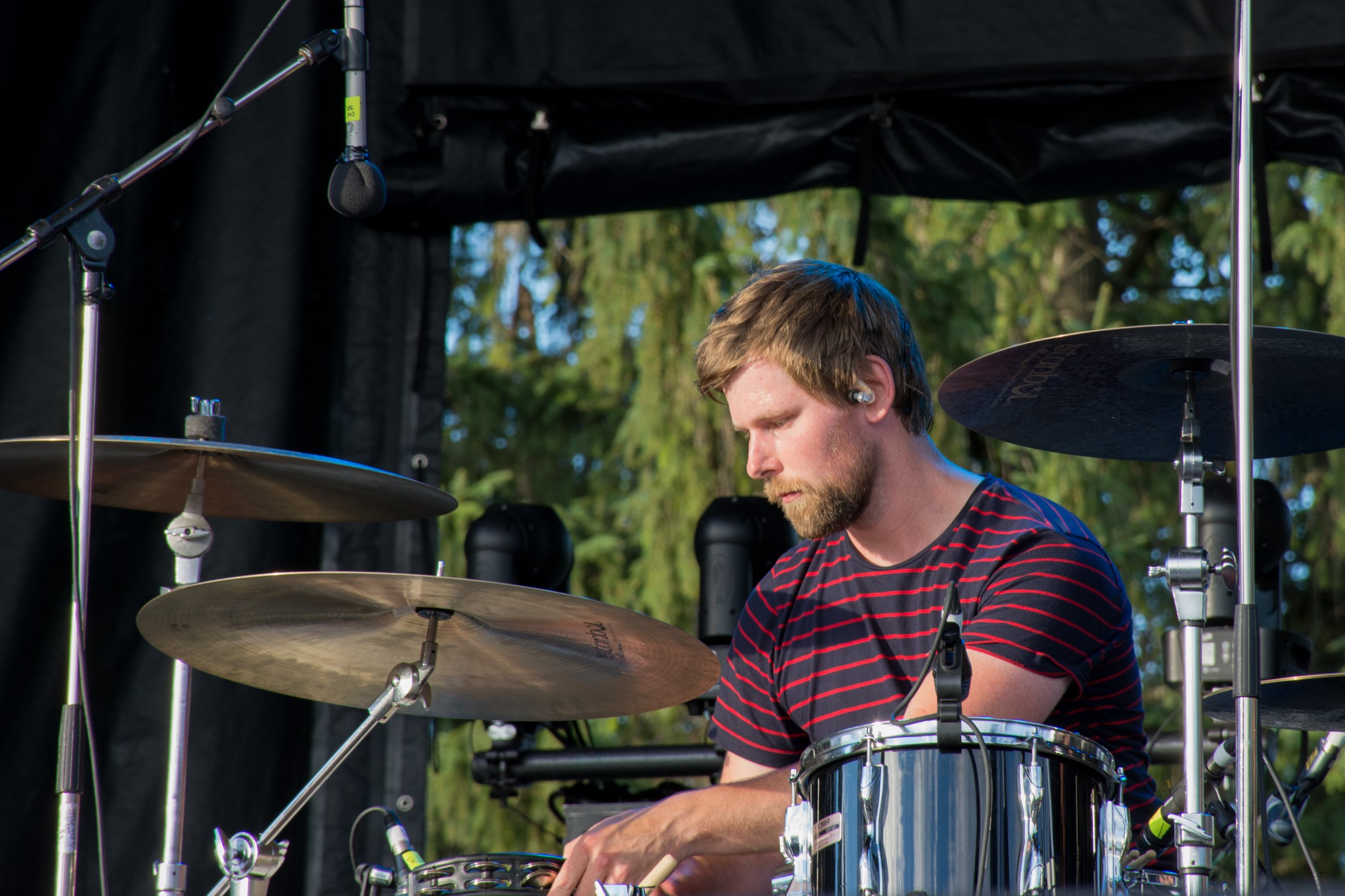 A man playing a drum set outdoors on a stage with trees in the background.