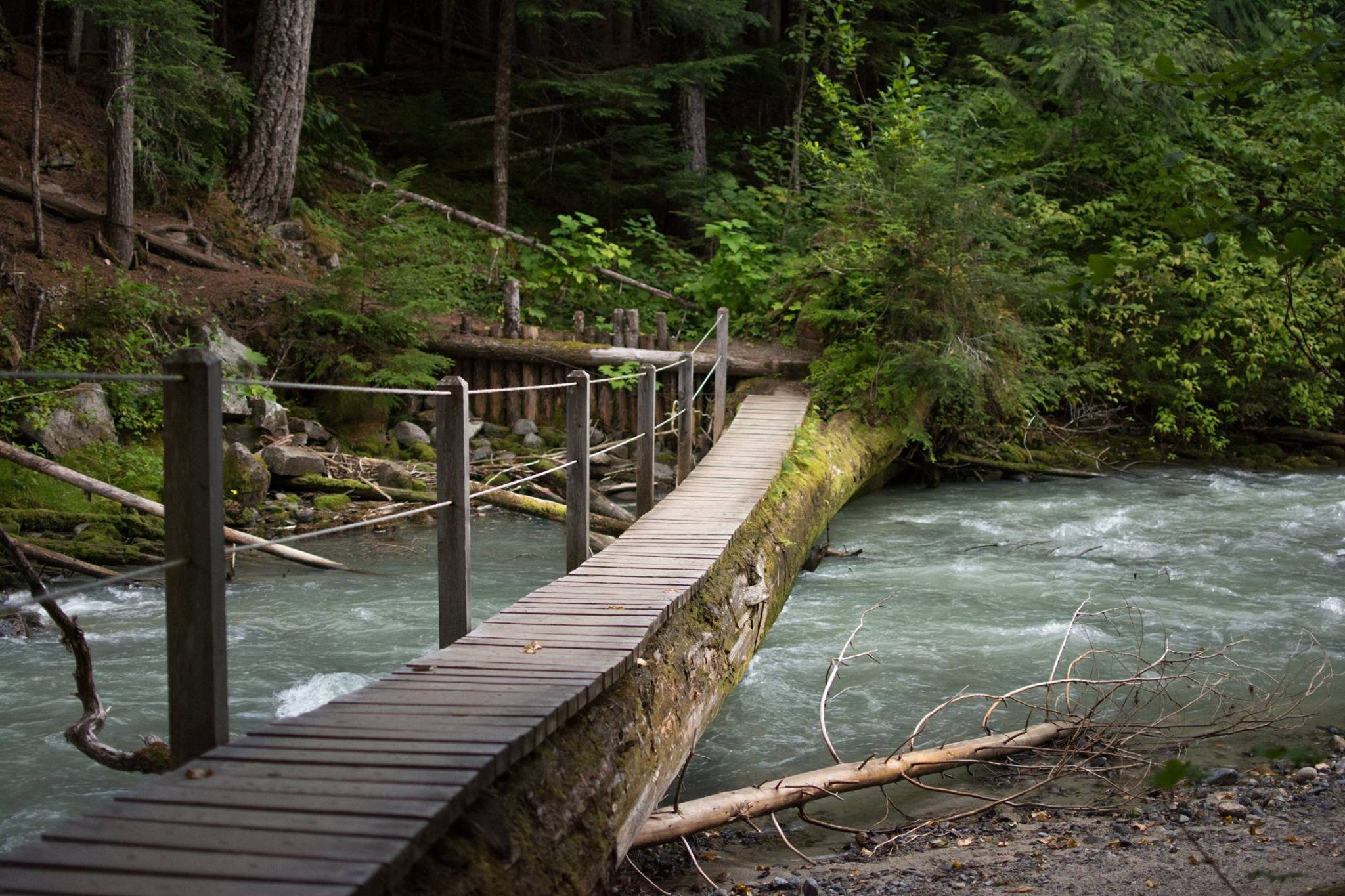 Wooden suspension bridge over a rushing river in a forested area.
