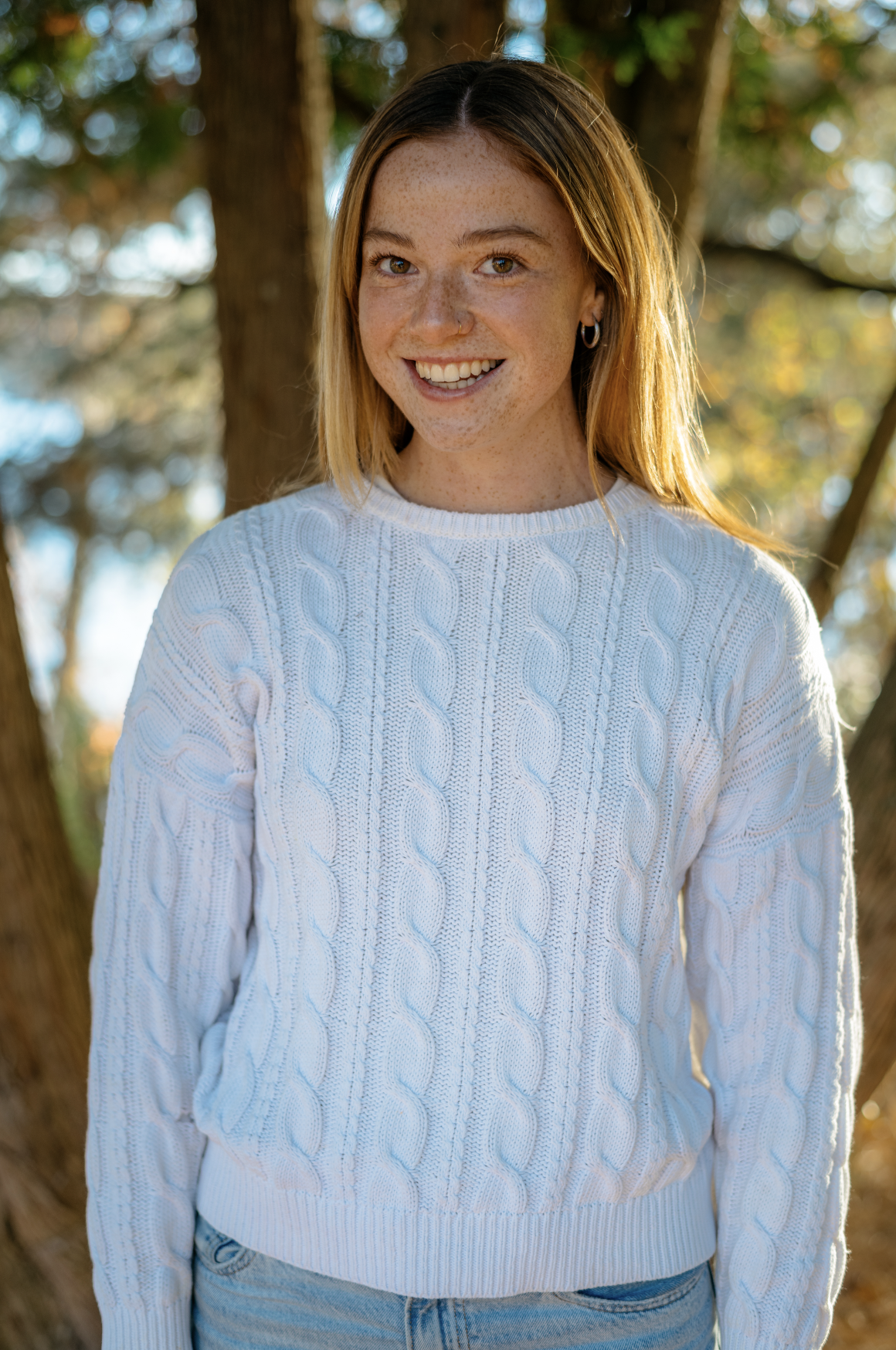 A young woman with red hair, freckles, and hoop earrings smiling outdoors in front of trees, wearing a white cable-knit sweater.