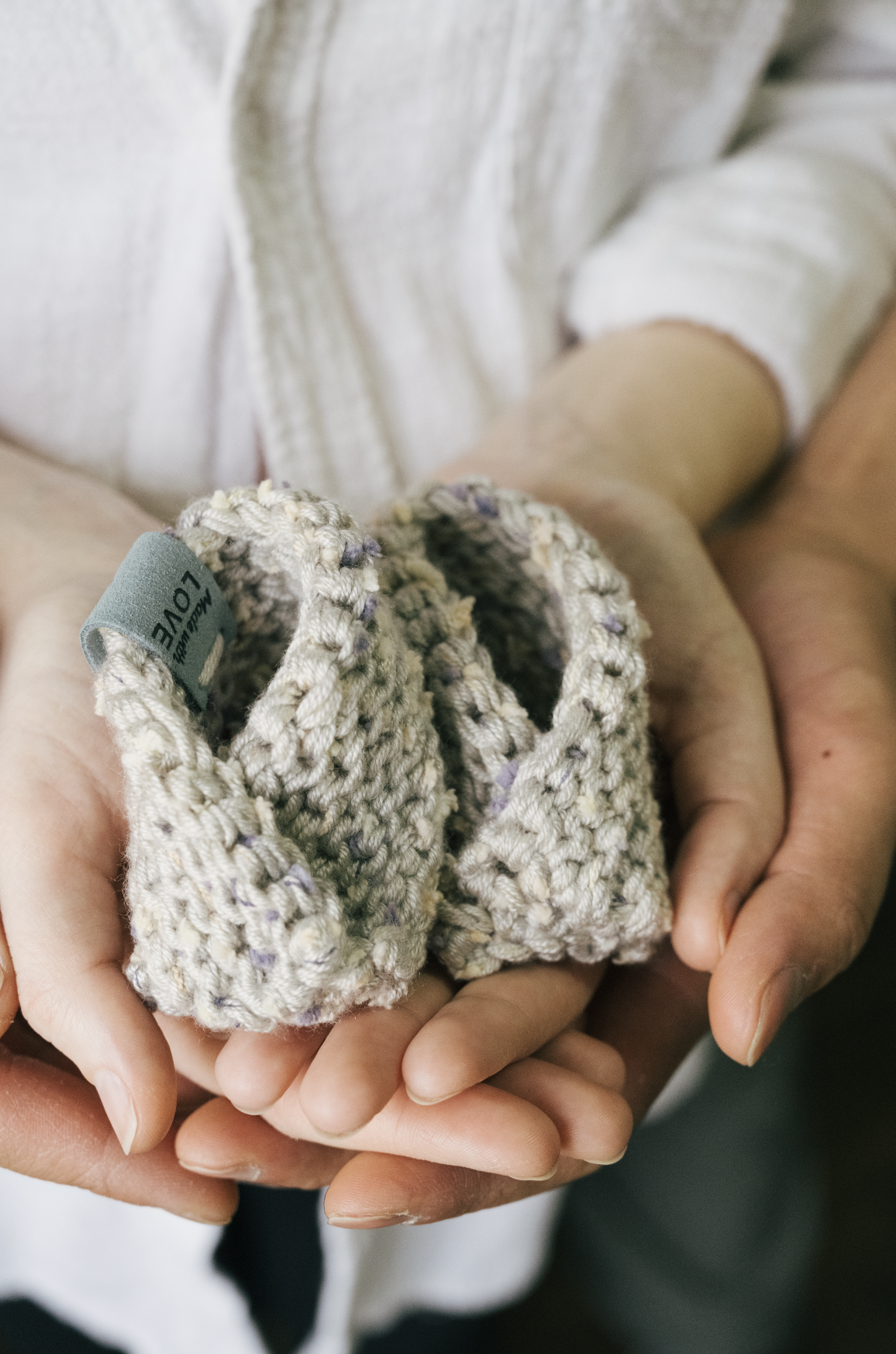 Close-up of a pair of tiny knitted baby booties being held in adult hands, with a person's body in the background.