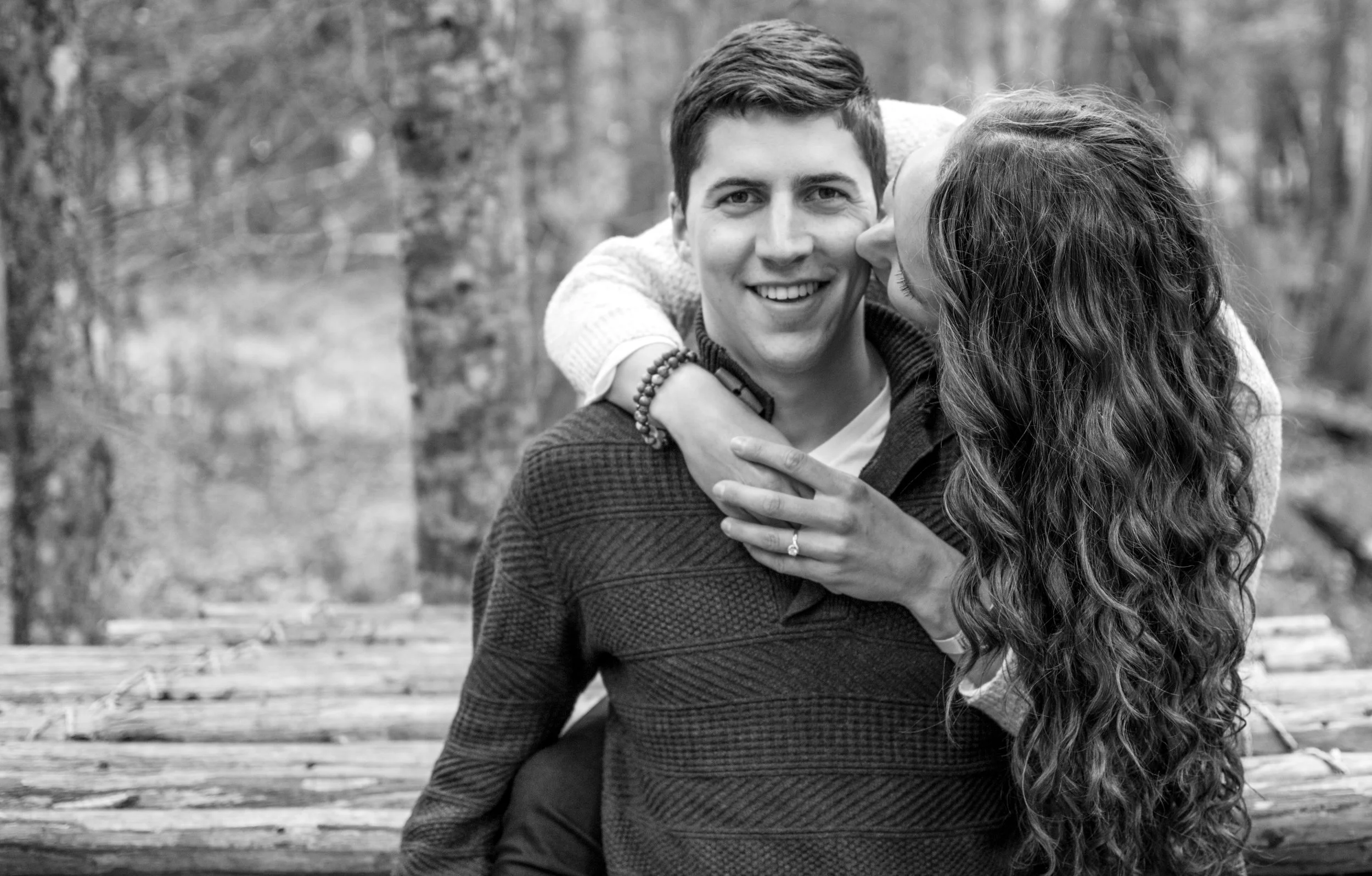 A young couple smiling, with the woman kissing the man's cheek, outdoors in a forest setting.