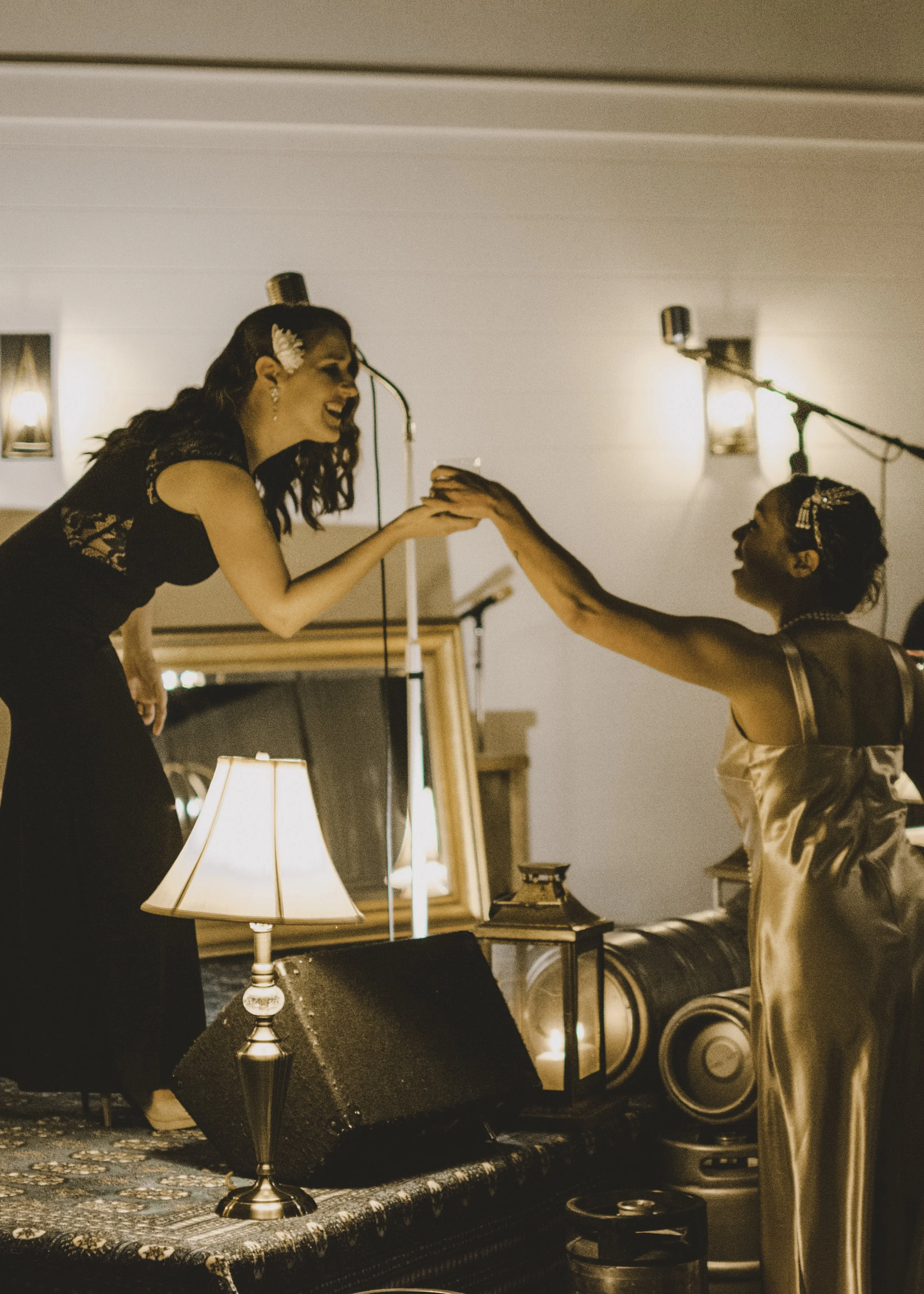 Two women at a celebration or wedding, with one woman on a stage reaching out to high-five the other woman in a satin dress. The scene is warmly lit with lamps and lanterns, and there is a mirror and speakers in the background.
