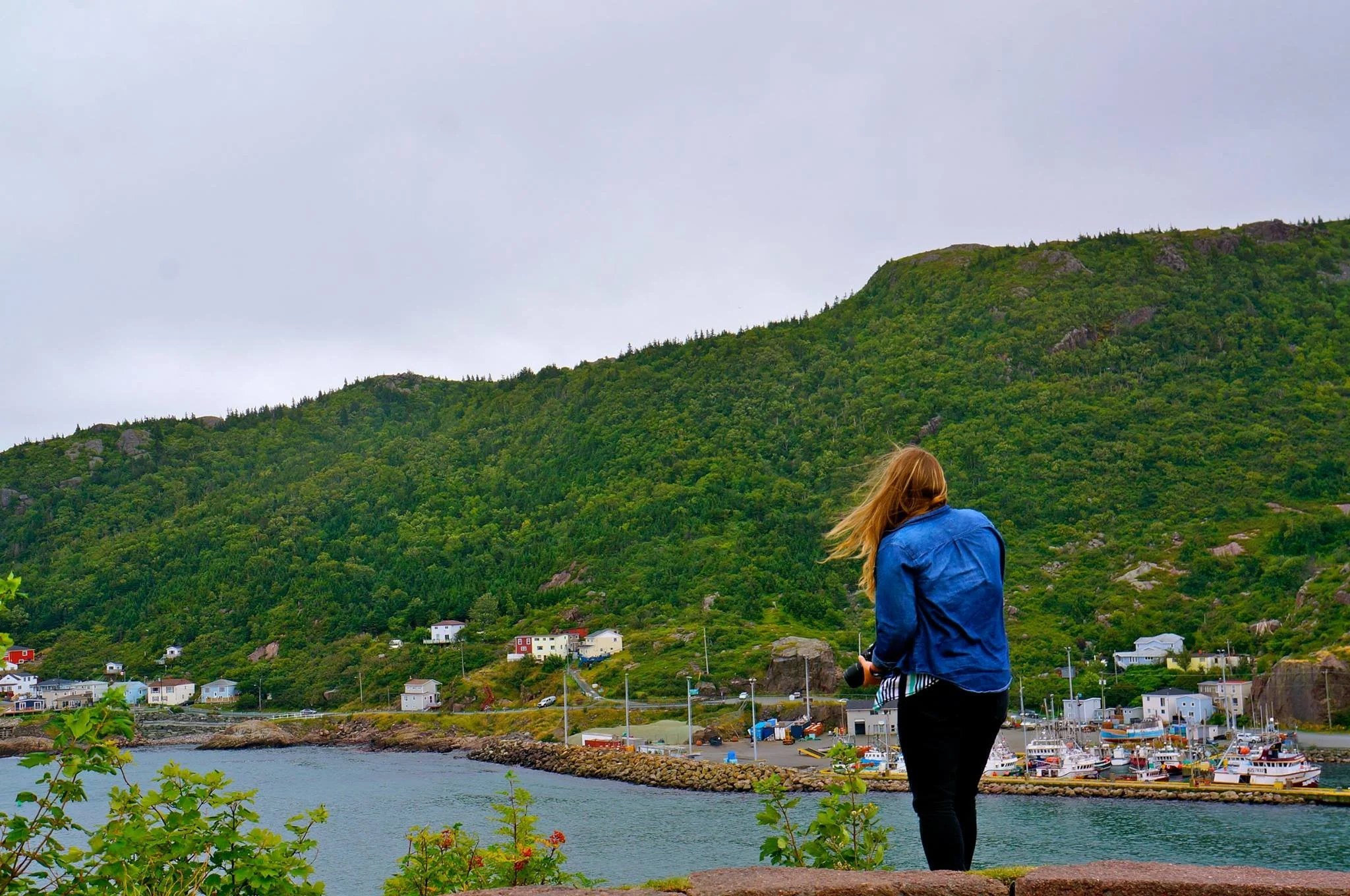 Woman with long hair in a blue jacket holding a camera, standing on rocks near a harbor with boats, overlooking a green hillside and small houses under cloudy sky.