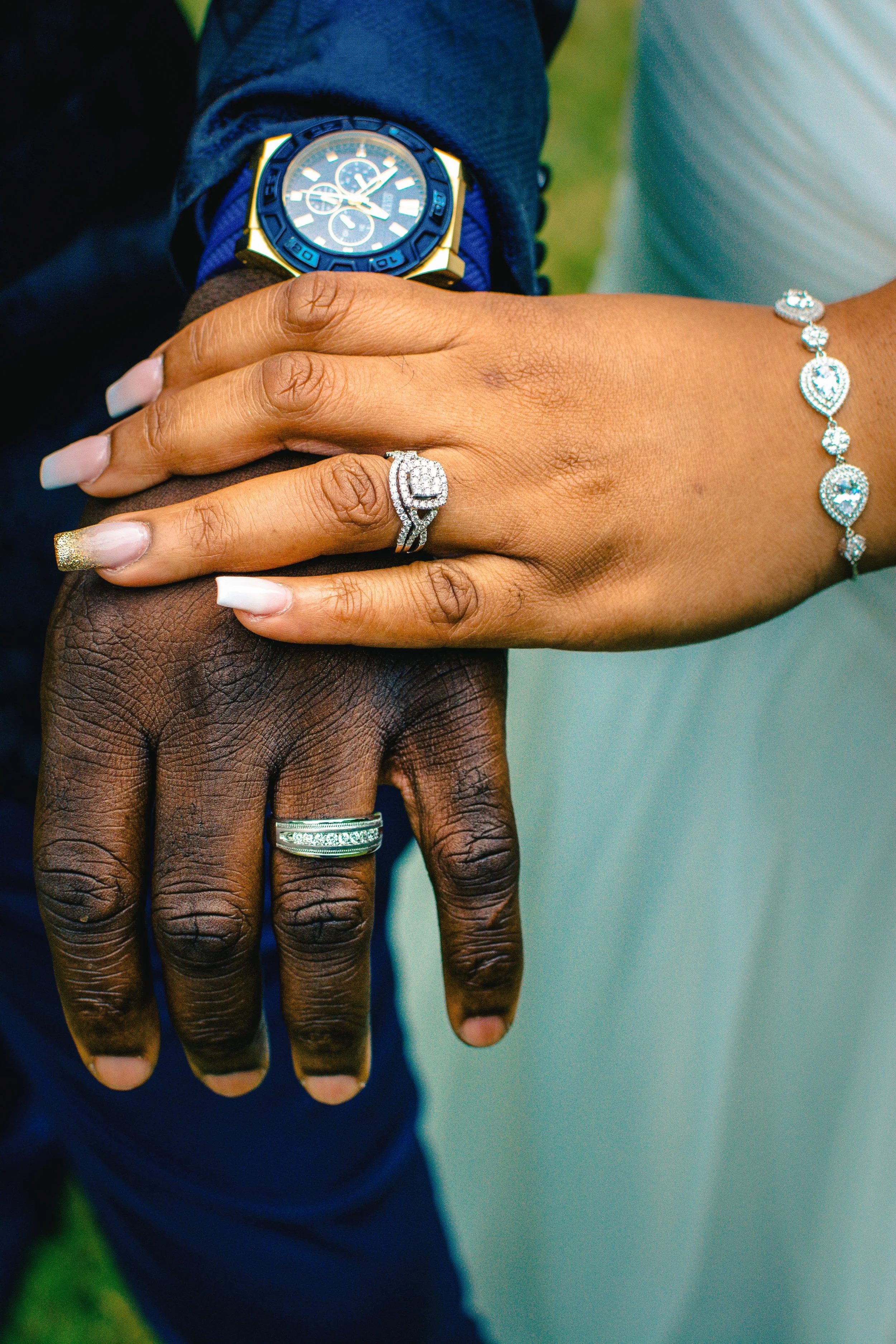 Close-up of a dark-skinned man's hand with two rings, and a light-skinned woman's hand with a diamond engagement ring and a diamond bracelet, resting on top of his hand.