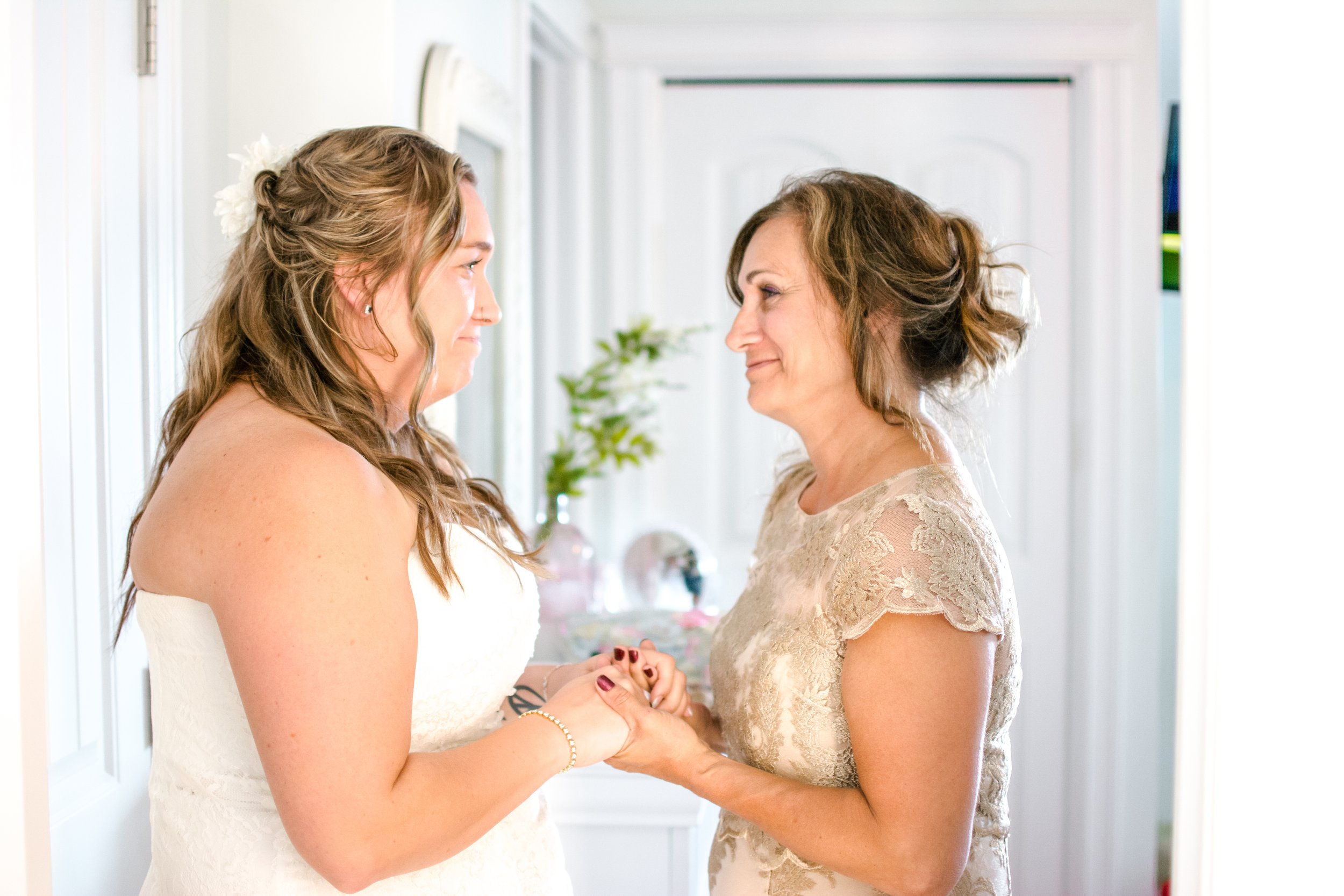 A bride and her mother holding hands and smiling at each other in a brightly lit room.