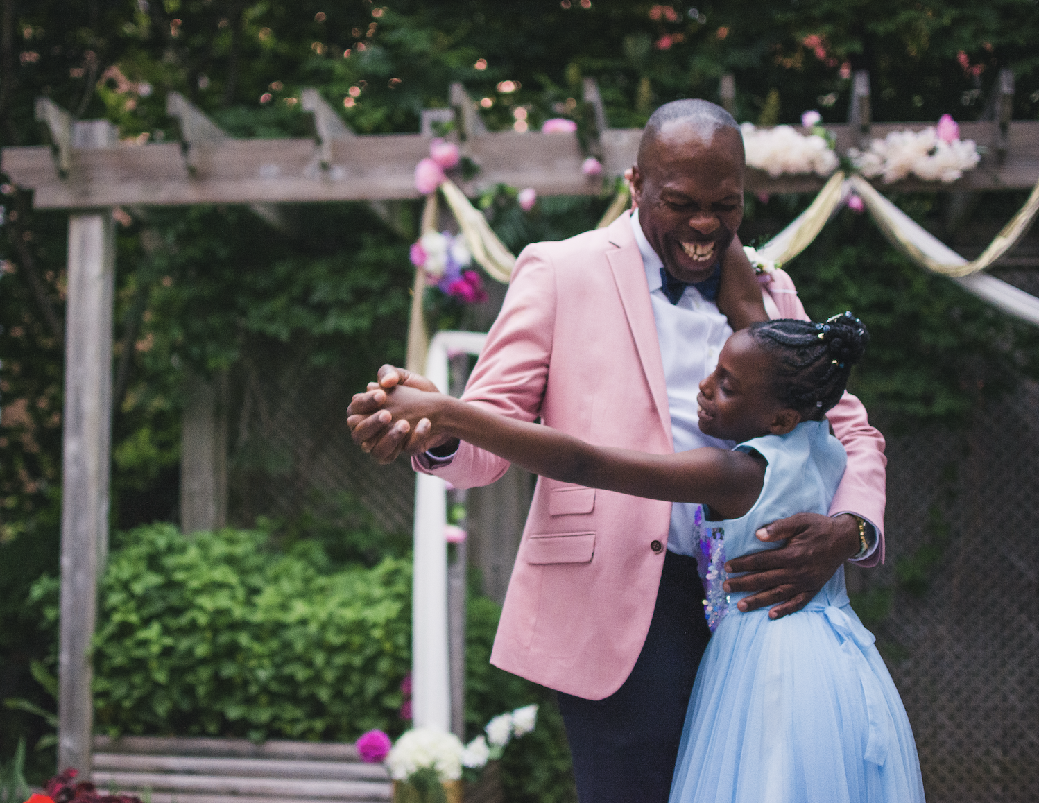 A man and young girl are dancing together in a garden. The man is wearing a light pink blazer, a white shirt, and a blue bow tie, while the girl is dressed in a light blue dress with her hair styled in braids. They are smiling and enjoying a moment of dance, with decorated flowers and drapery in the background.