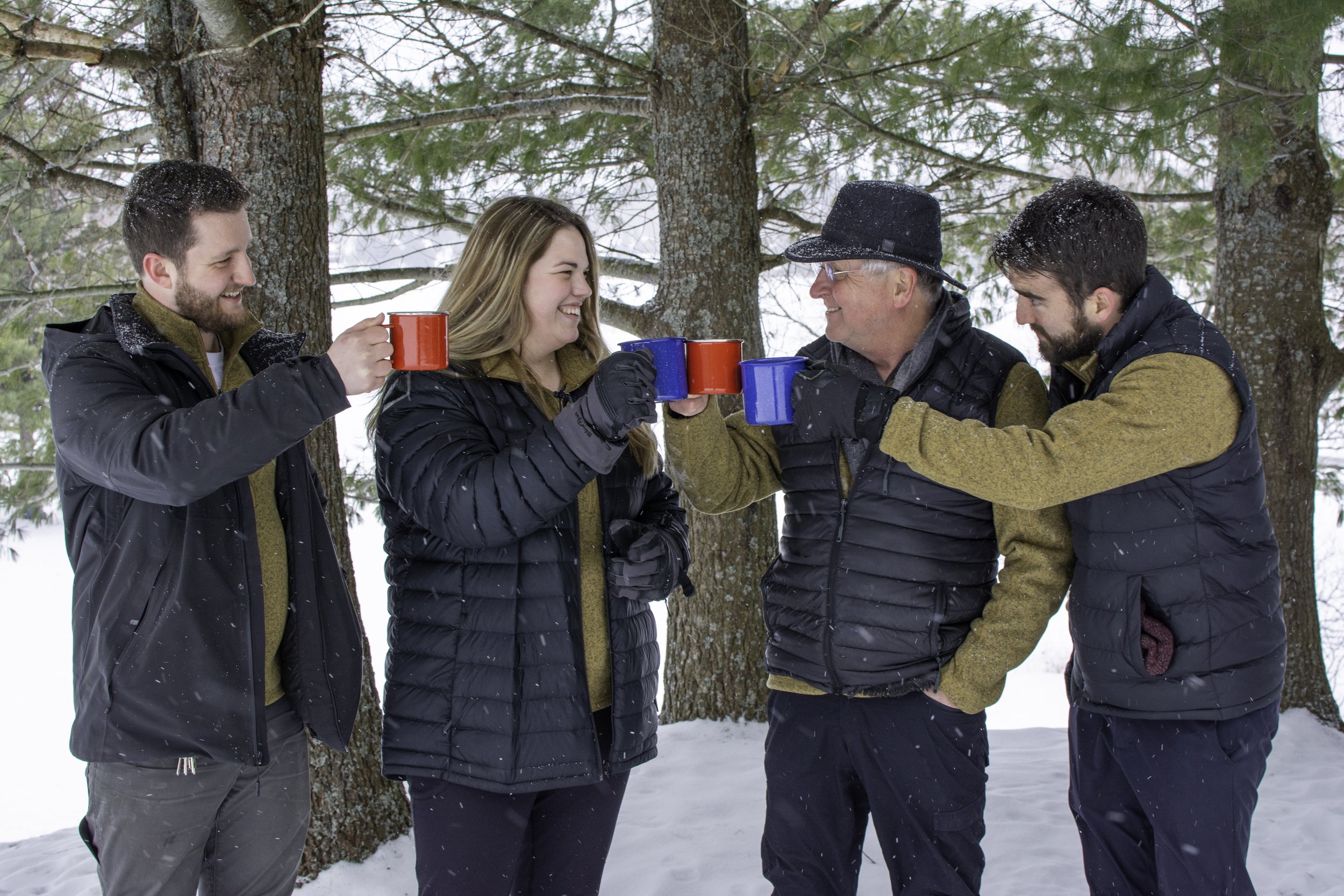 Four people standing outdoors in the snow, raising red and blue mugs in a toast, smiling, with trees in the background.