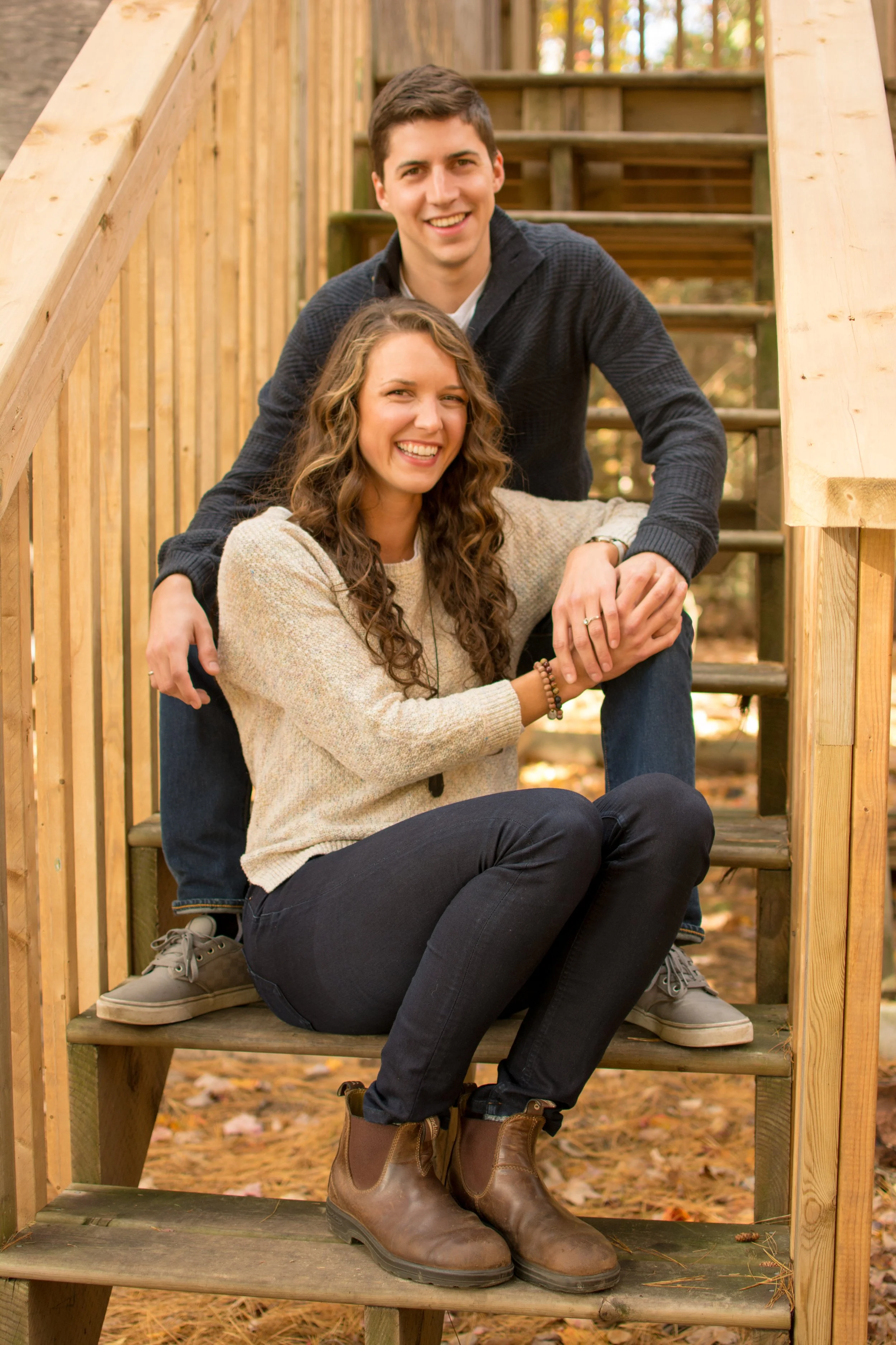 A young man and woman smiling, sitting on wooden stairs outdoors in fall, surrounded by autumn leaves.
