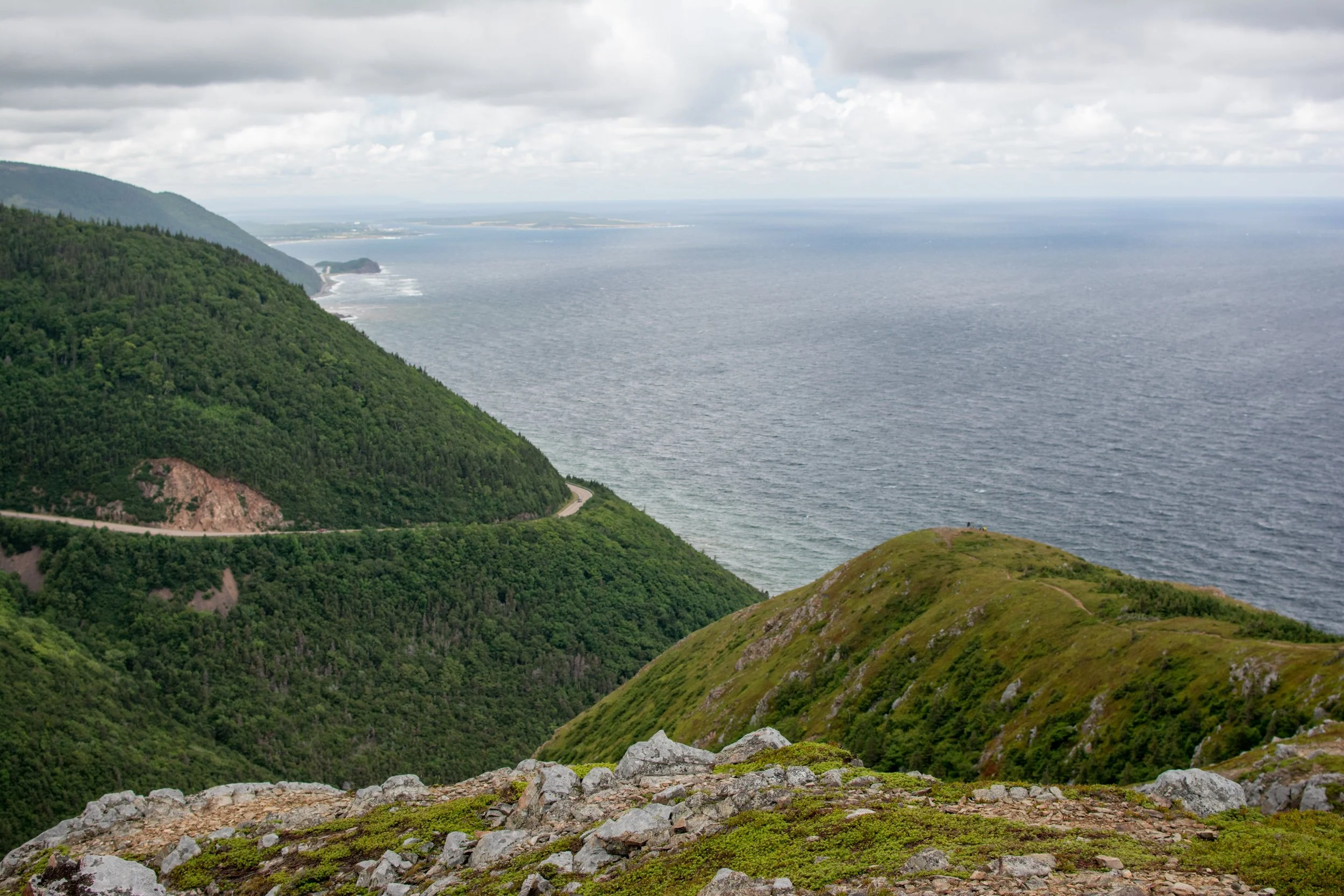 A scenic view of lush green hills with a winding road, overlooking the ocean under a cloudy sky.