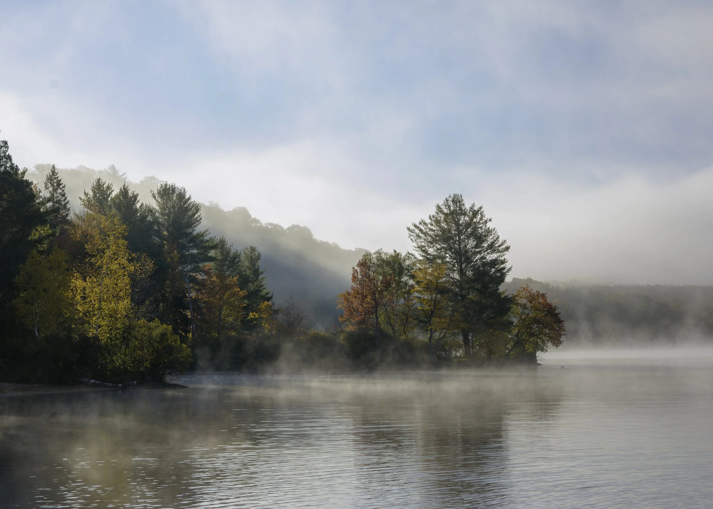 A peaceful river scene with fog rising over the water, surrounded by trees with autumn foliage, and a partly cloudy sky above.