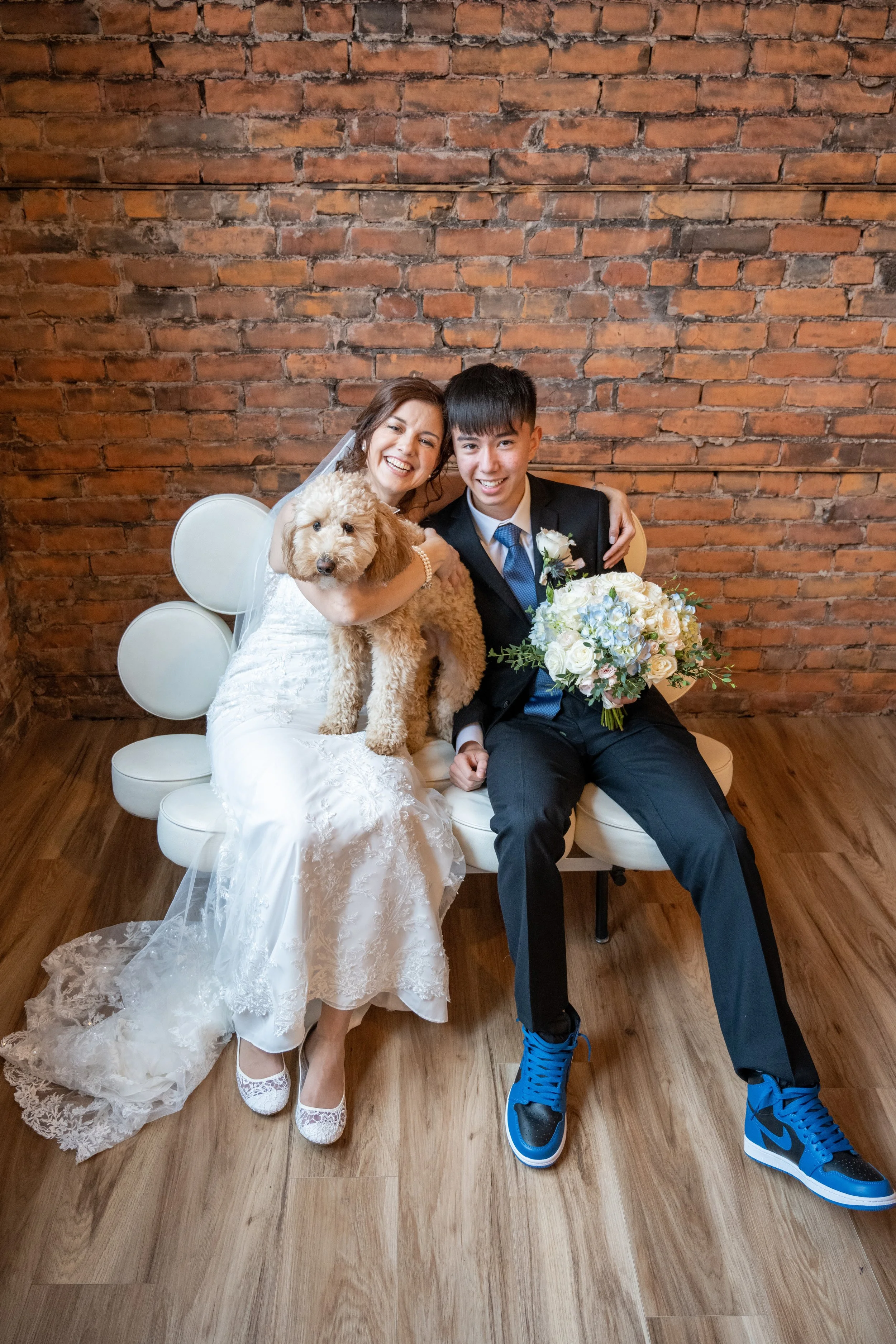 A bride and groom sitting on a white bench in front of a brick wall, smiling and holding a small fluffy dog and a bouquet of flowers.