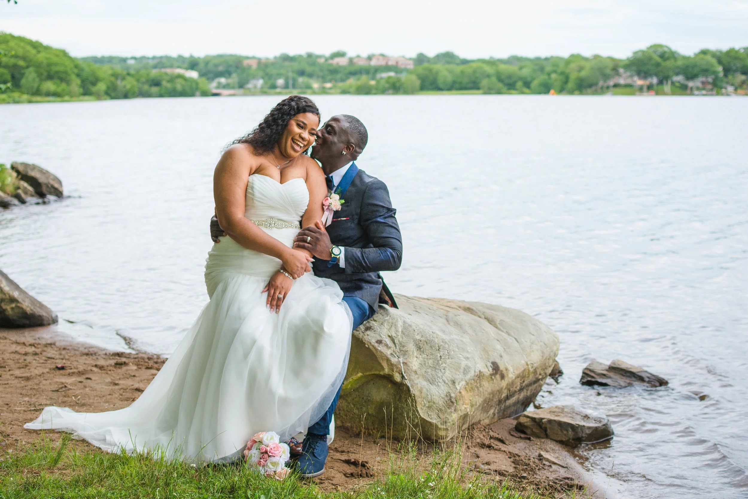 A newlywed couple sitting on a large rock at the edge of a lake, smiling and embracing, with greenery and houses in the background.