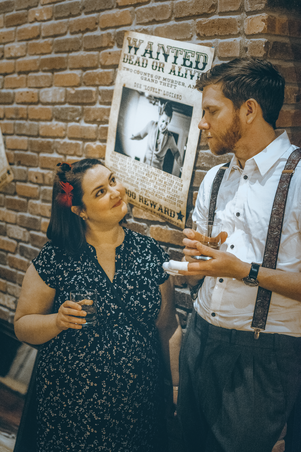 A woman and a man having a conversation at a bar, holding drinks, against a brick wall with a vintage wanted poster.