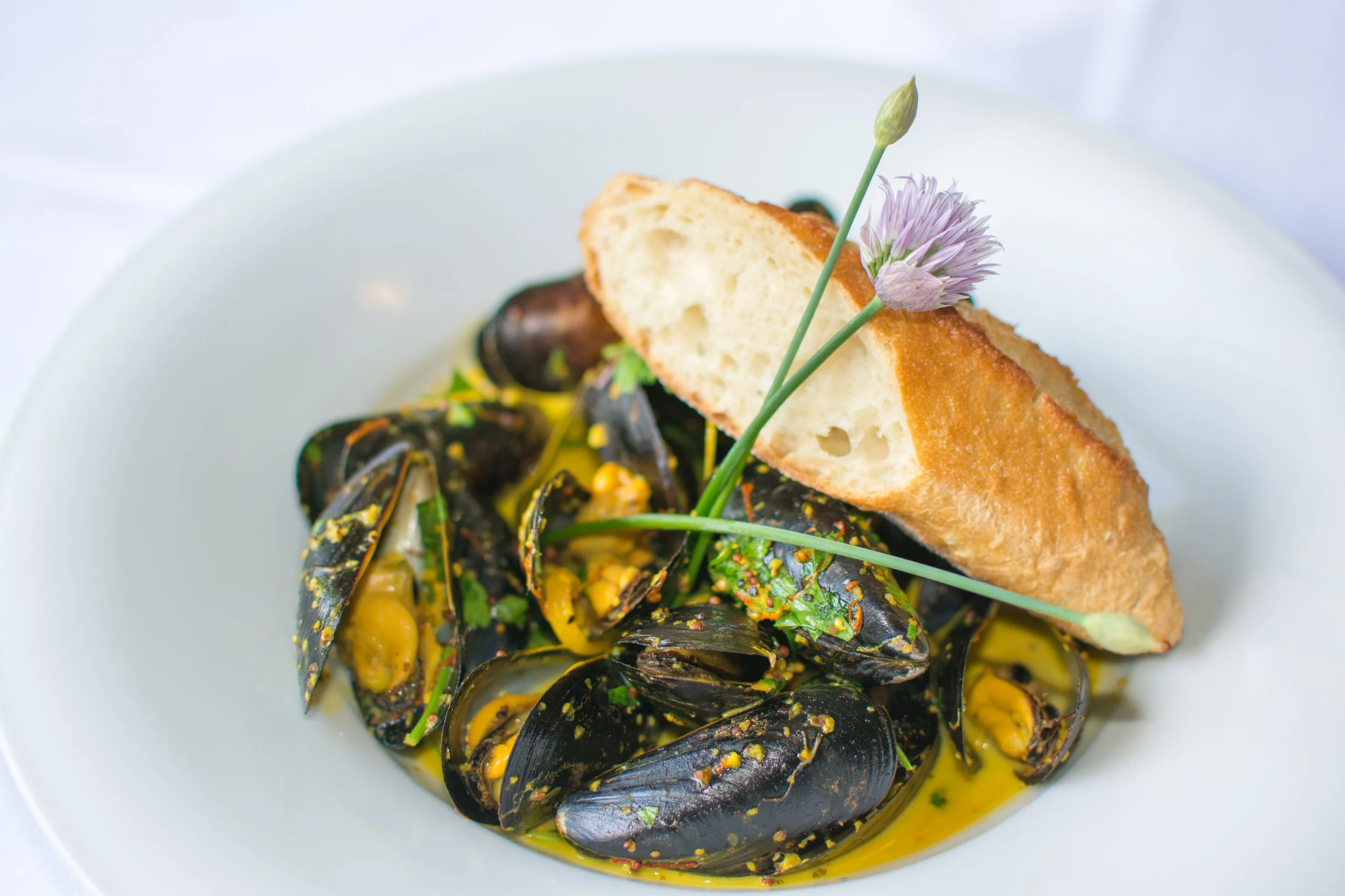 A white plate of mussels in yellow sauce, topped with a slice of baguette and garnished with a purple edible flower.