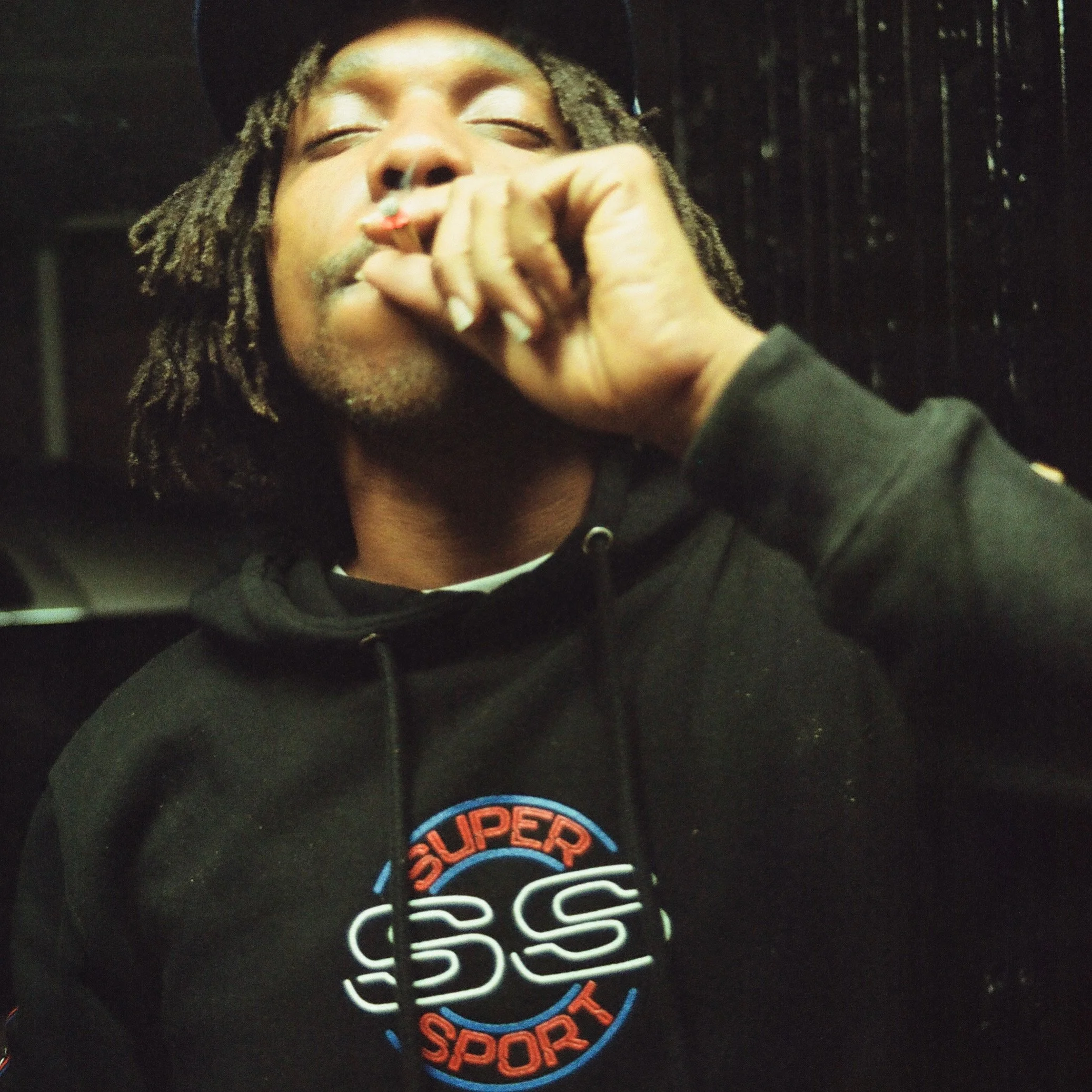 A person with dreadlocks smoking a cigarette, wearing a black hoodie with a colorful 'Super Sport' logo on it.