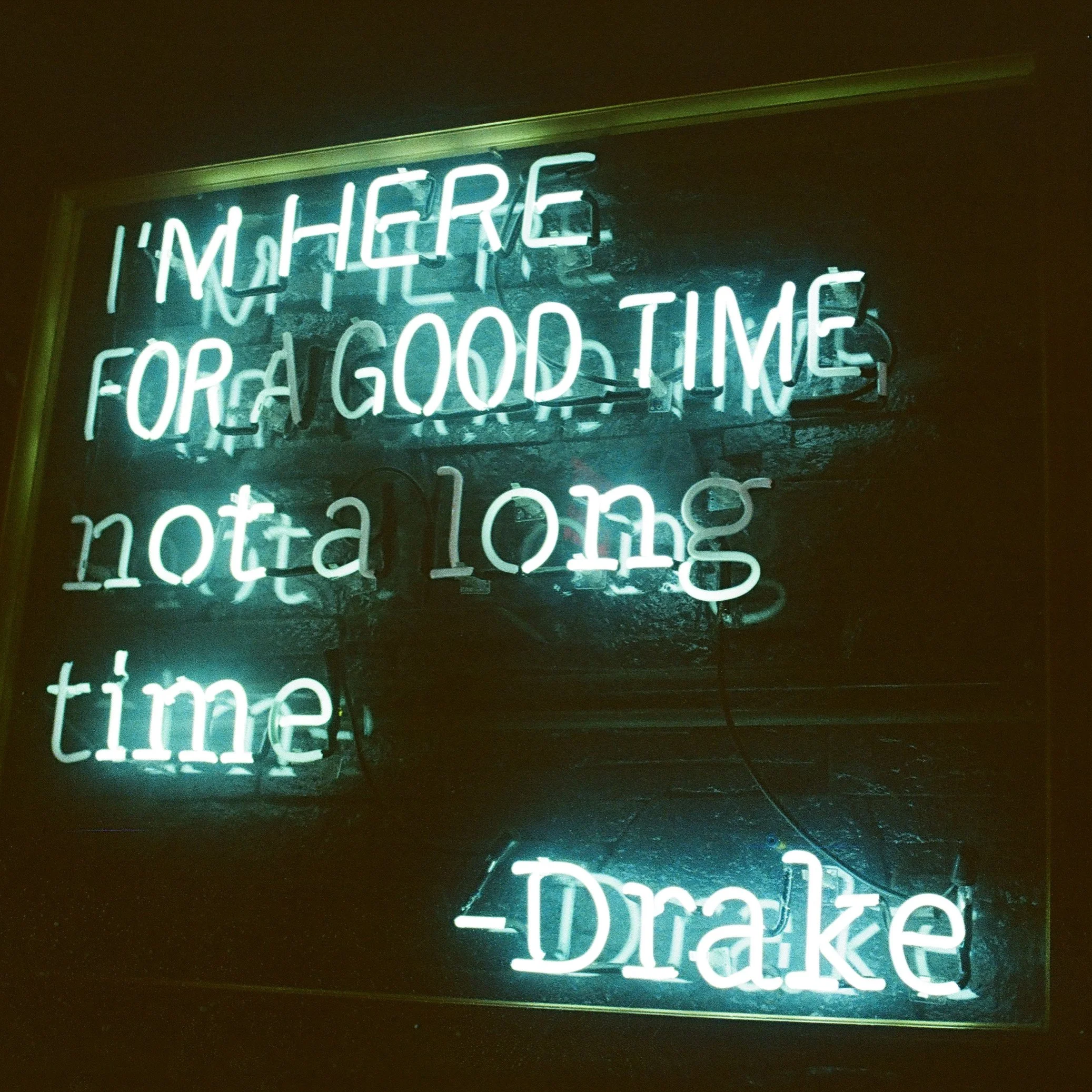 Neon sign with quote: 'I'm here for a good time, not a long time' attributed to Drake, illuminated in white and blue, mounted on a dark wall.