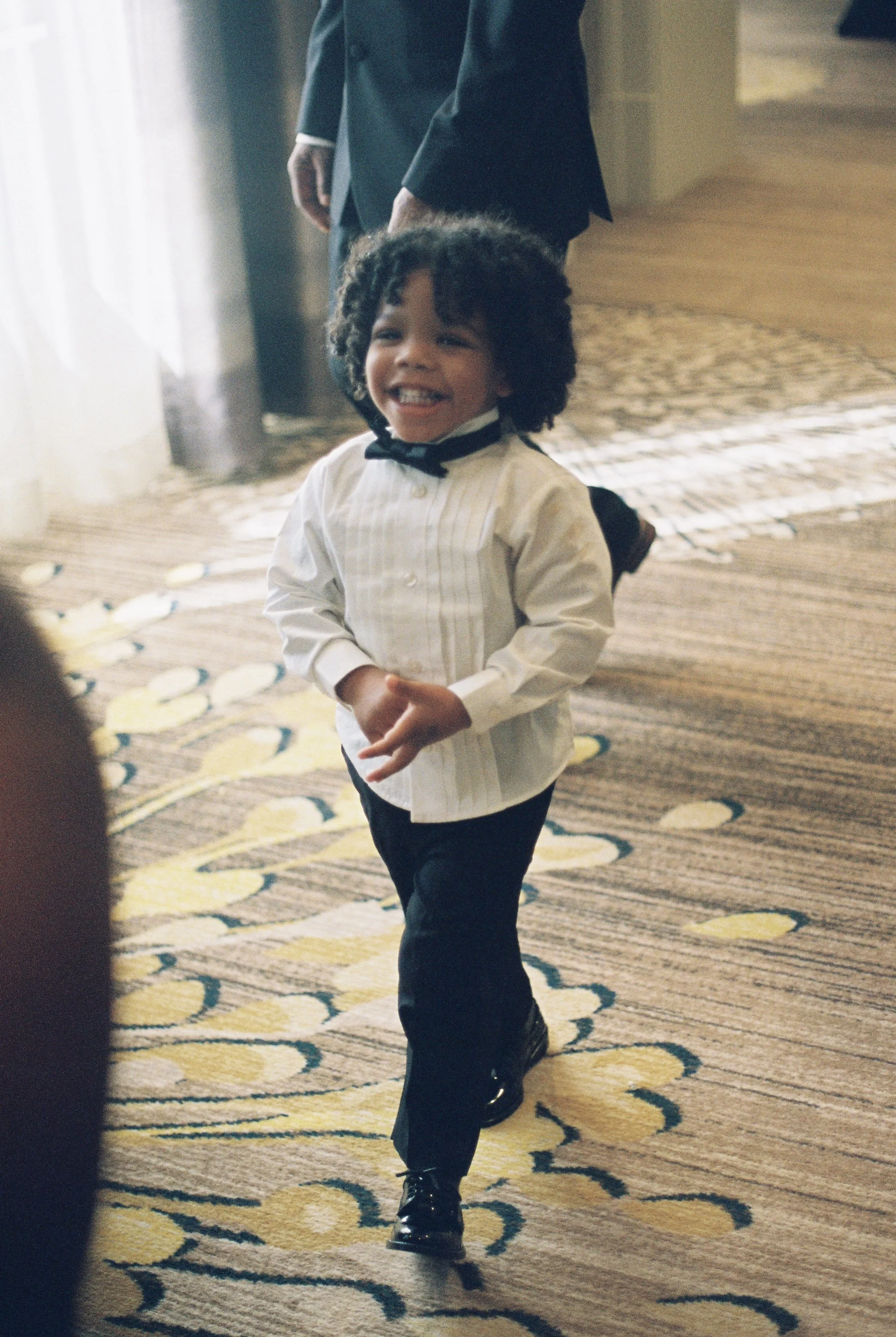 A young boy with curly hair smiling and walking on a patterned carpet, dressed in a white shirt and dark pants, with a man in a suit standing behind him.
