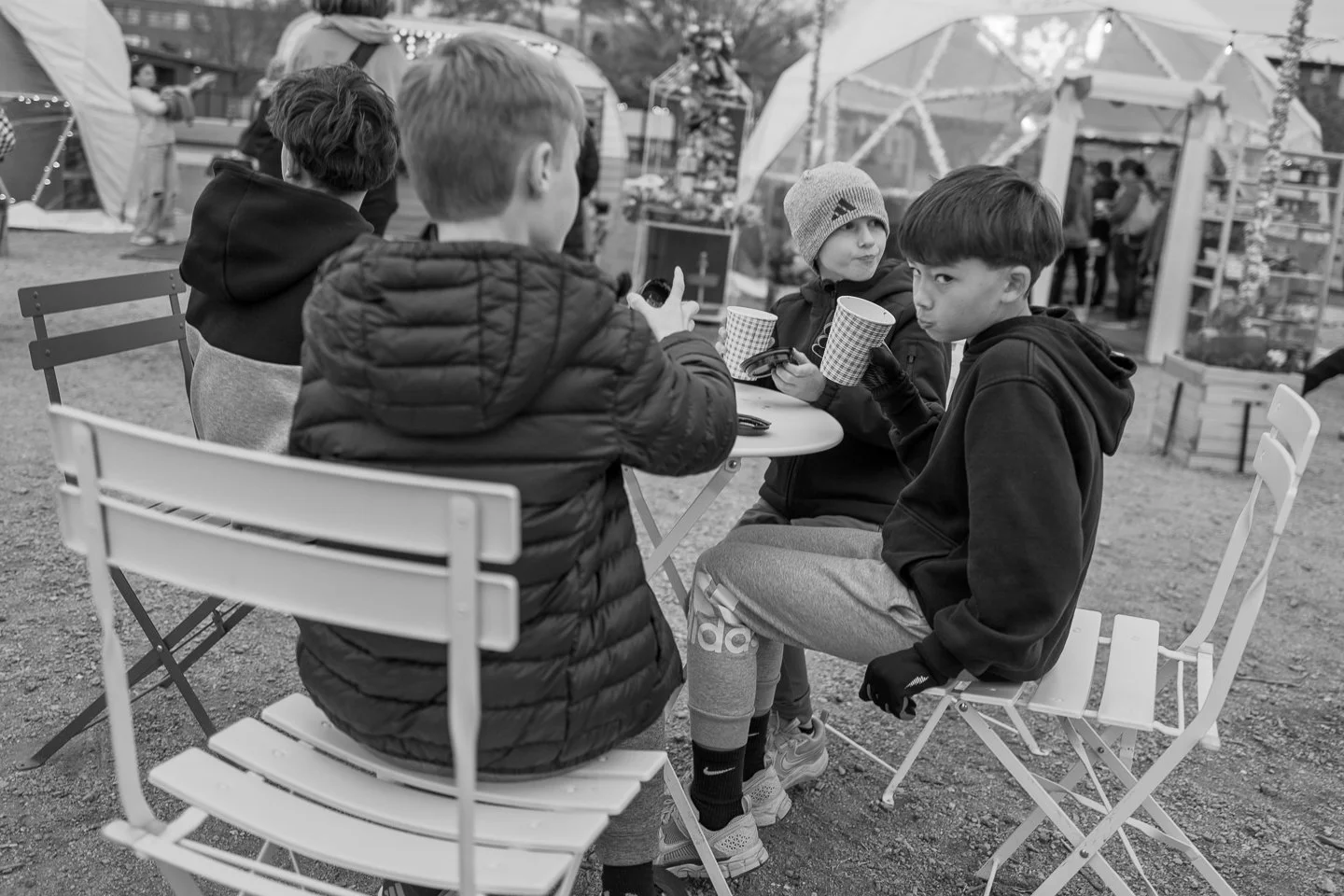 Four young boys sitting at an outdoor table, holding paper cups, with a festive market or fair in the background