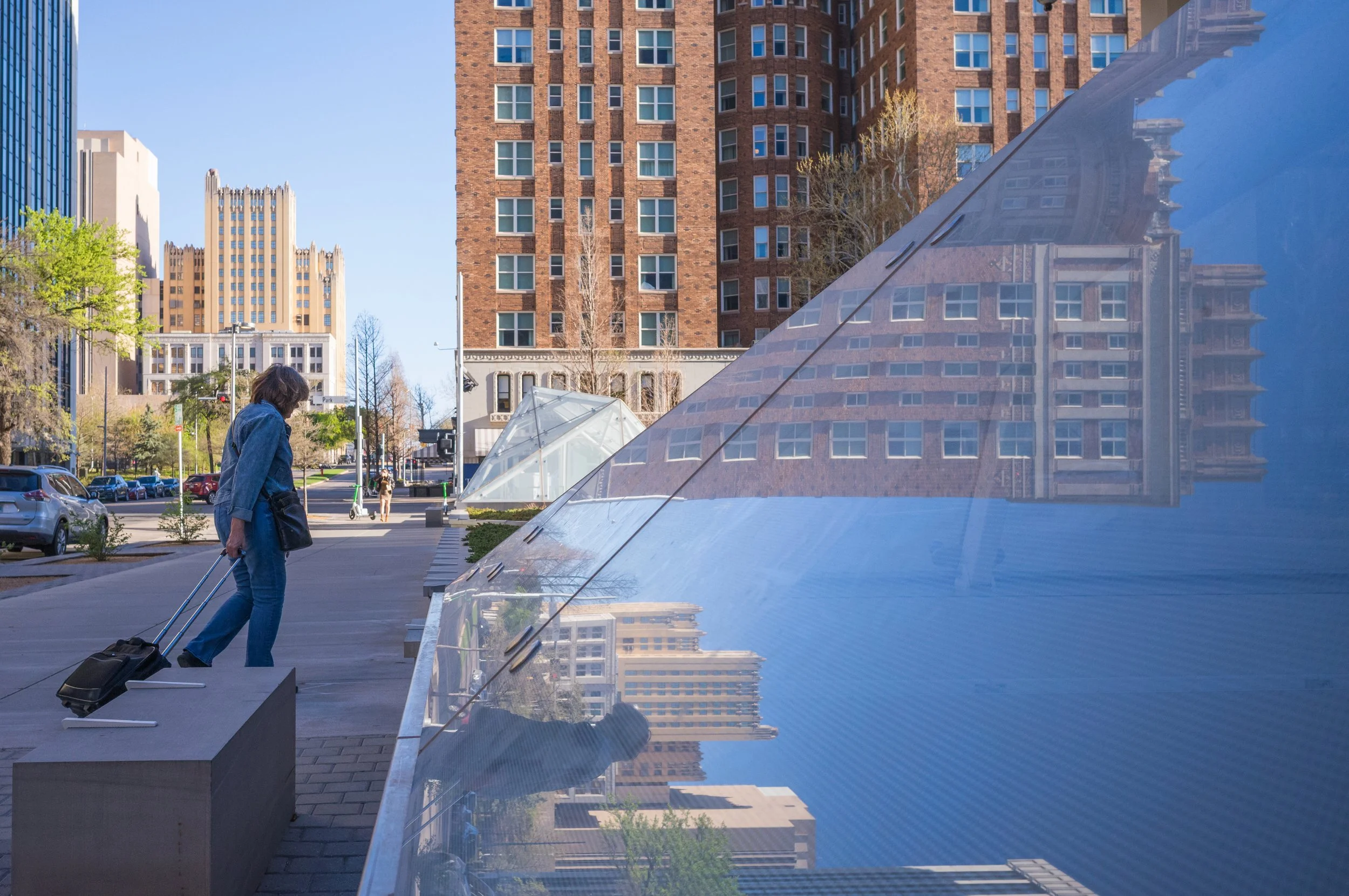 A person pulling a rolling suitcase walks past a reflective glass wall during daytime in an urban area with tall buildings.